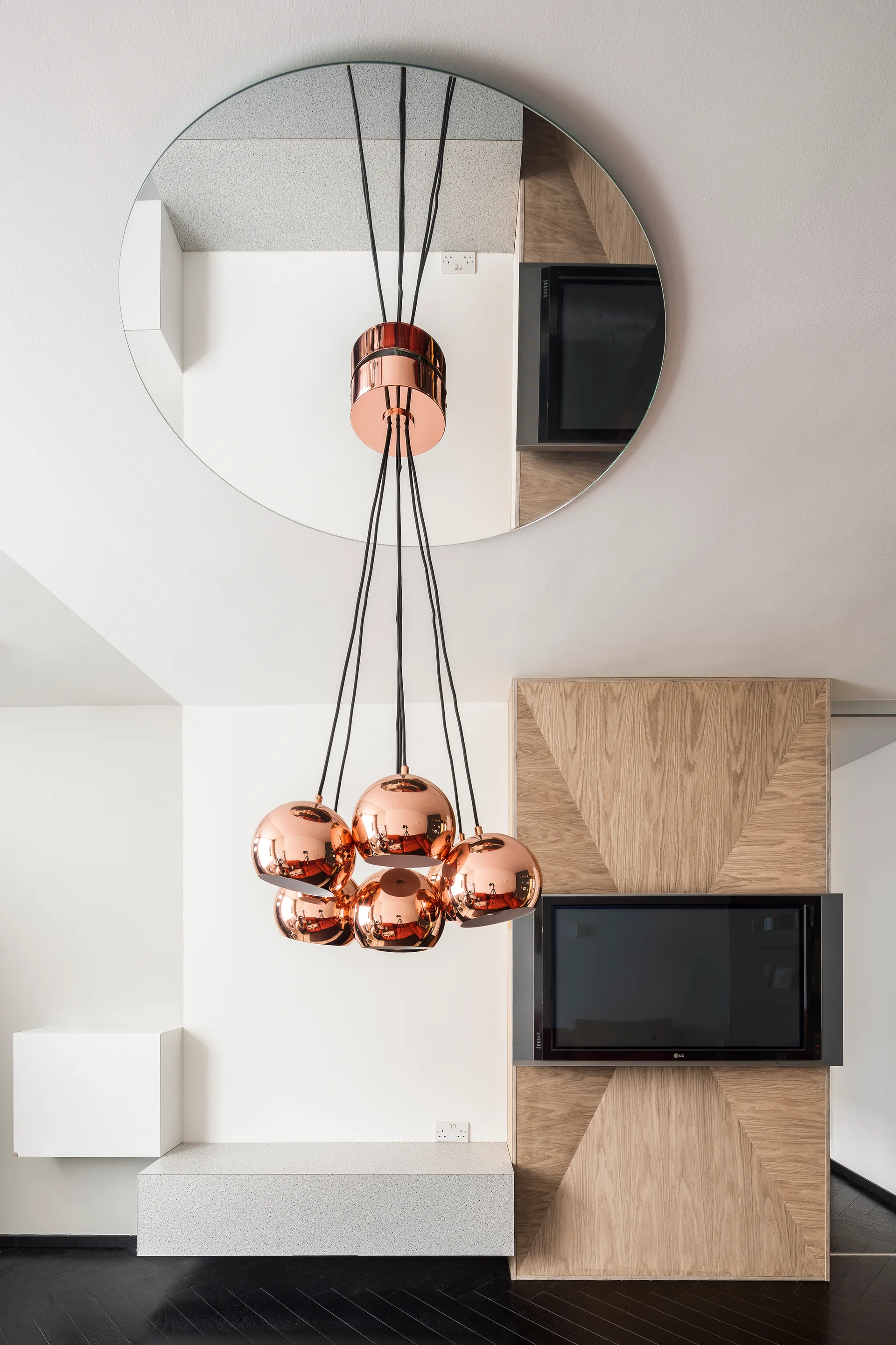 A modern living room detail showing a copper sphere chandelier hanging beneath a round ceiling mirror next to a wood-paneled TV wall.