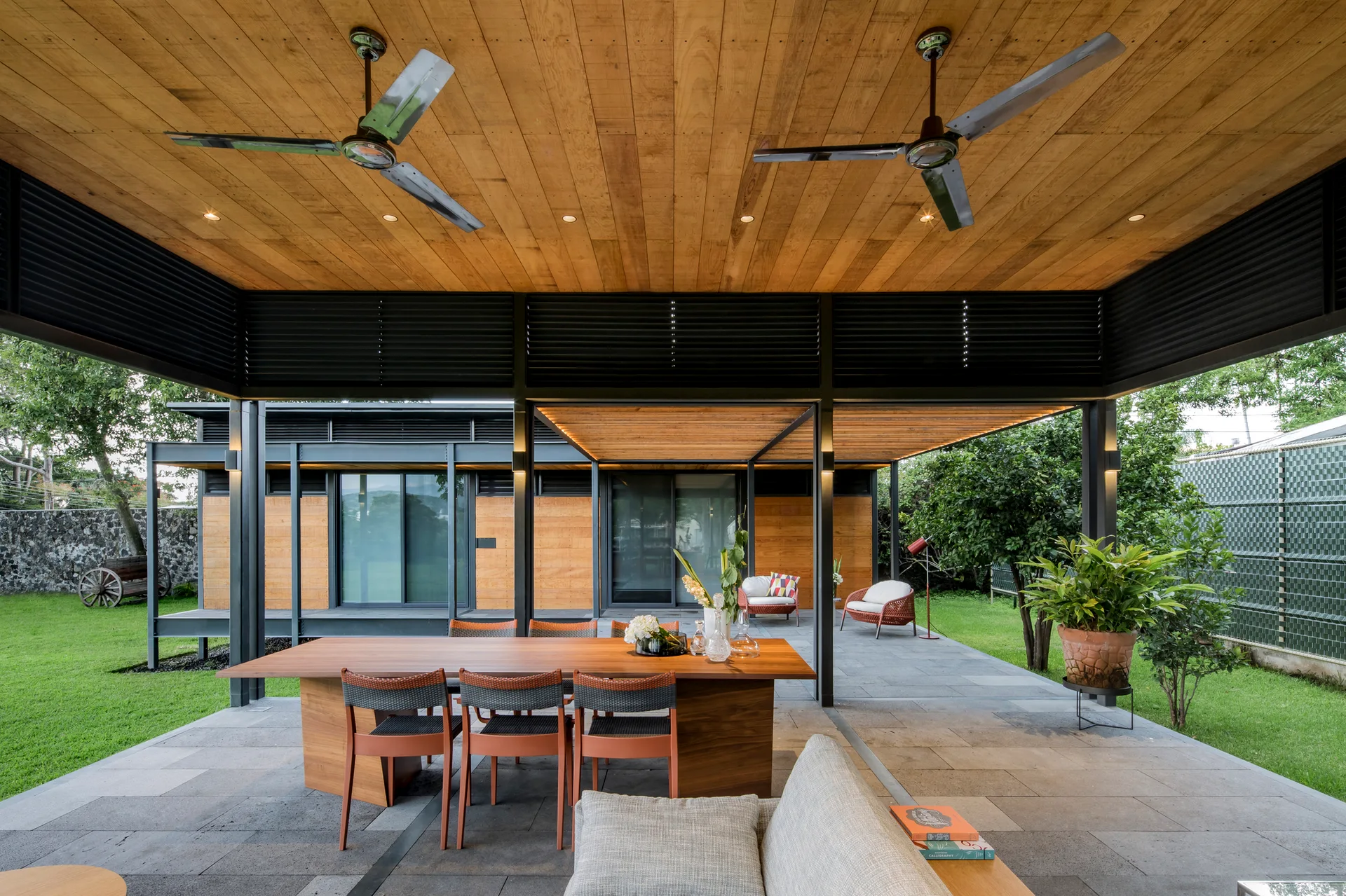 A covered outdoor patio featuring a wooden dining table and chairs, stone tile flooring, and a warm wood plank ceiling with ceiling fans.