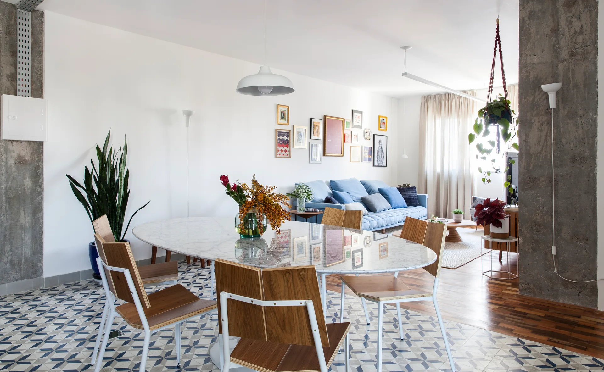 An open-plan dining space features an oval marble table, wood-and-metal chairs, and blue geometric tile flooring alongside an exposed concrete column.
