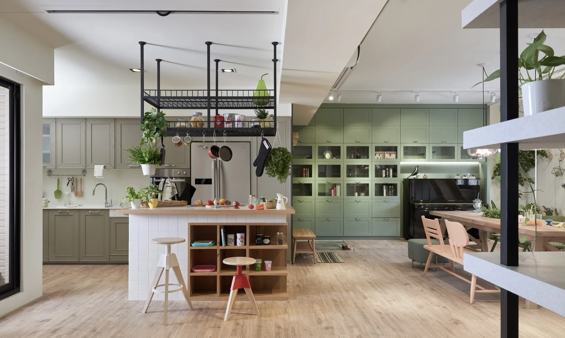 An open kitchen featuring sage green cabinets, a wood-topped white tiled island with stools, a hanging black metal pot rack, and dining table.