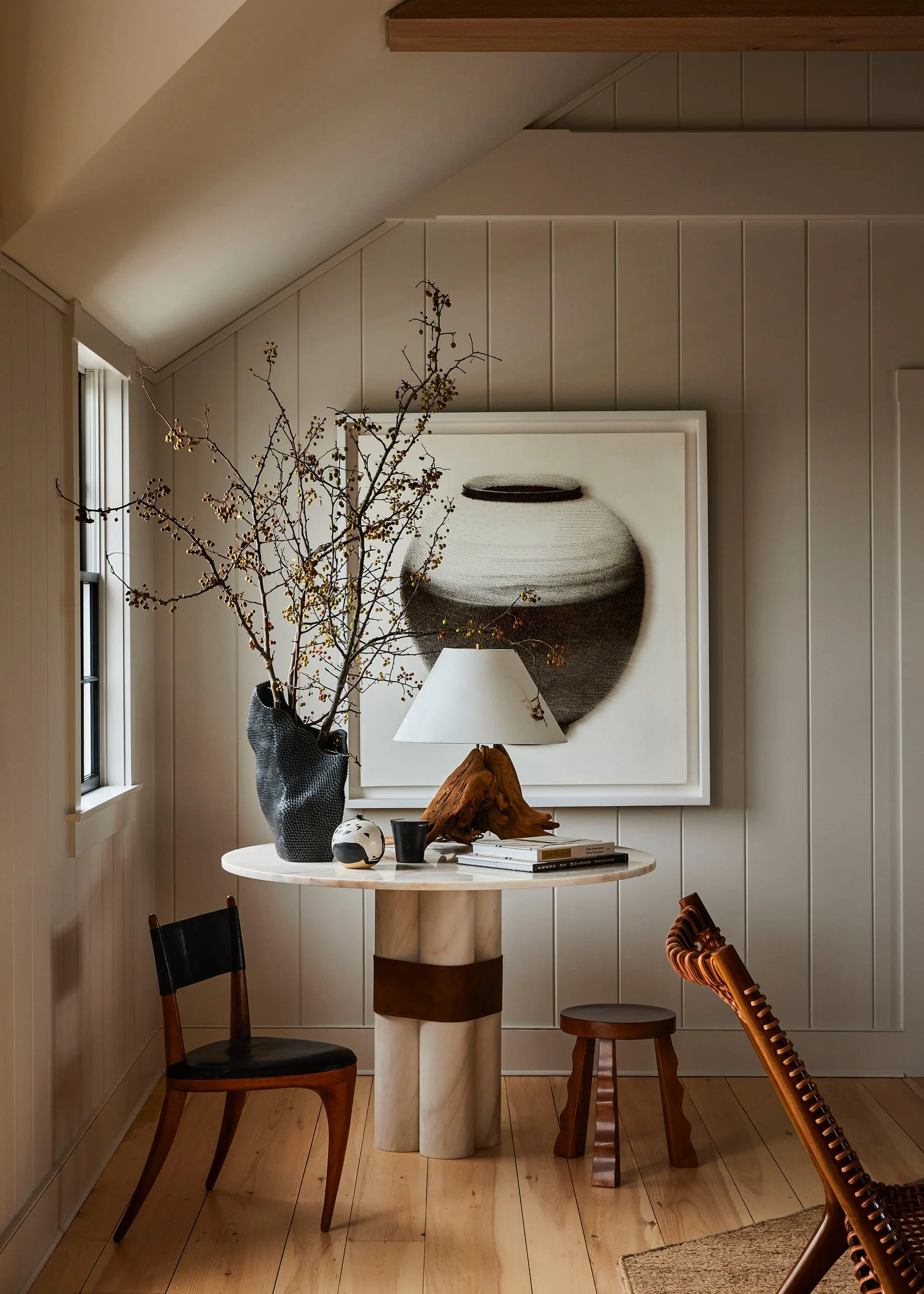 A round stone pedestal table with a rustic driftwood lamp and a black vase of branches stands against a vertical paneled wall.