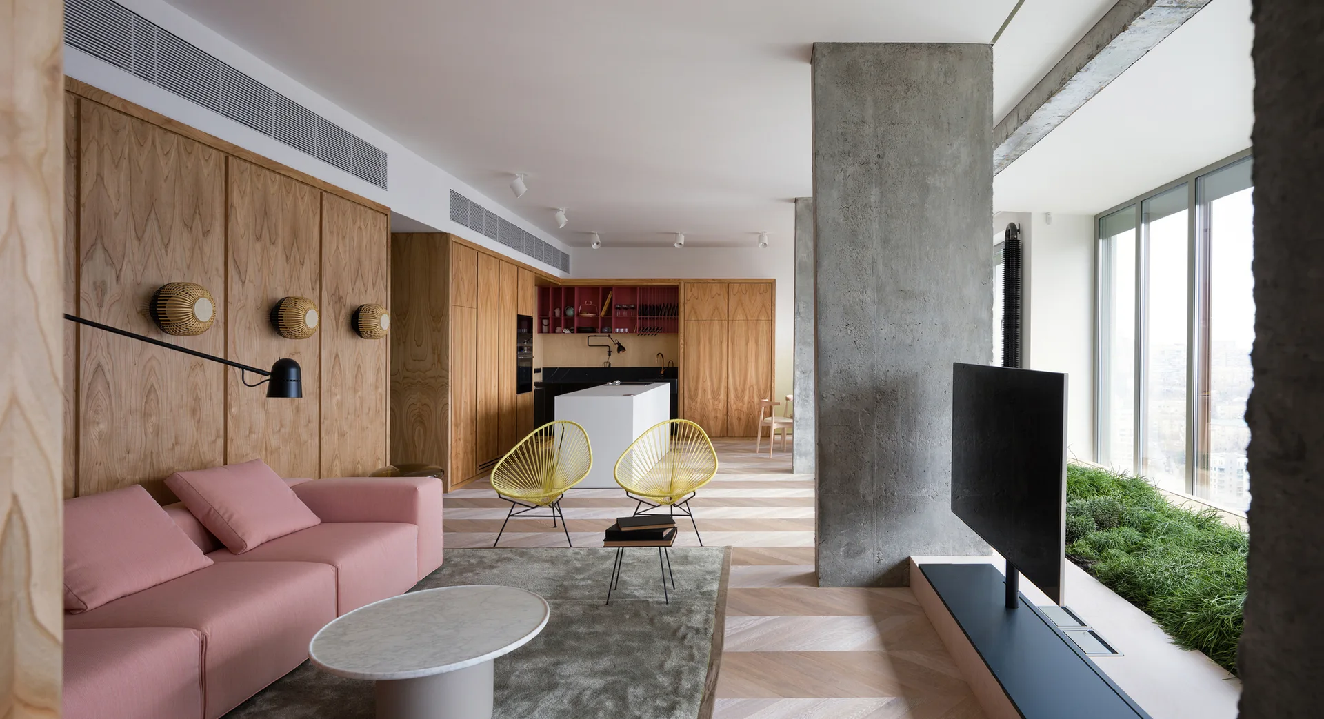 A modern open-plan living room featuring a pink modular sofa, yellow wire chairs, a raw concrete column, and wood-paneled walls leading to a kitchen.