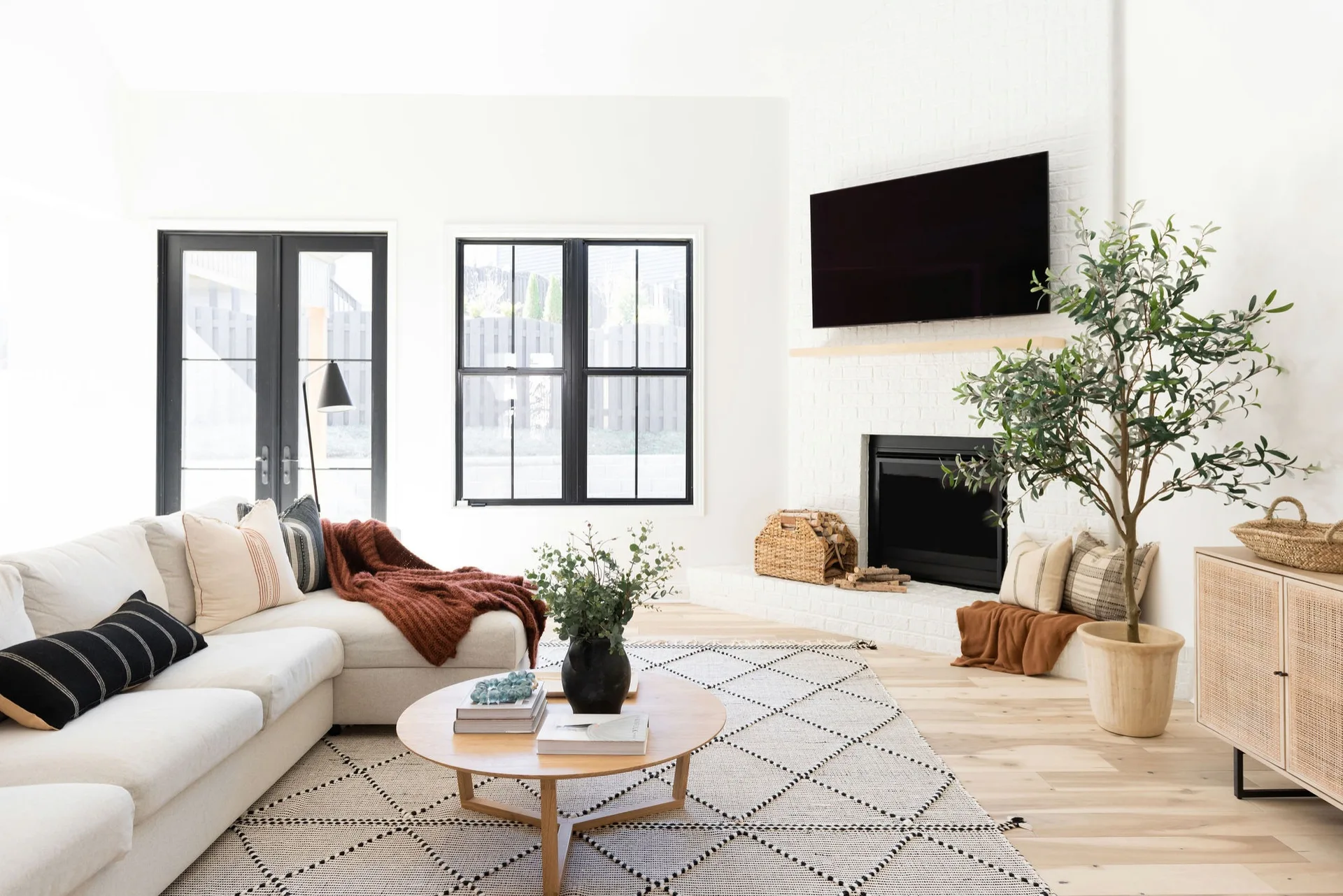A bright living room features a cream sectional sofa, a round wooden coffee table on a geometric rug, and a white painted brick fireplace.