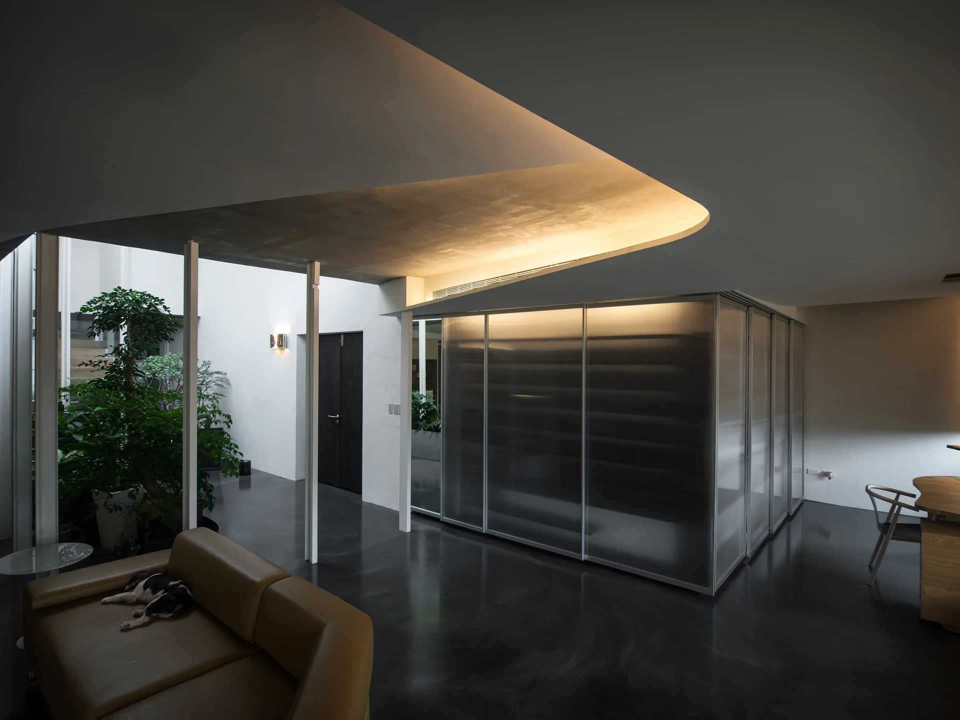 A modernist living room features a tan leather sofa, a curved illuminated ceiling soffit, a ribbed glass room divider, and dark polished floors.