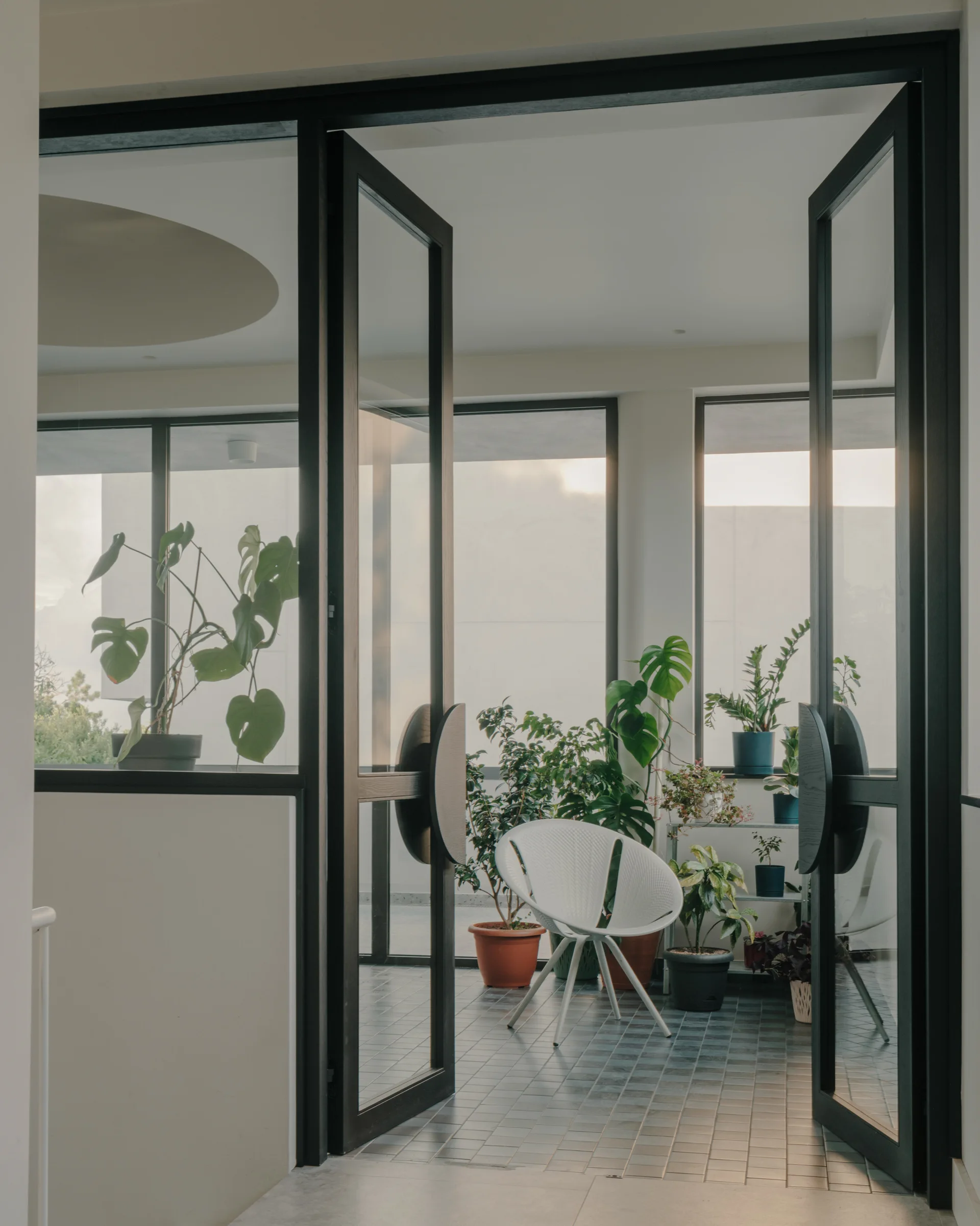 Open black-framed glass French doors lead into a sunlit tiled patio featuring a white modern chair surrounded by various potted green plants.