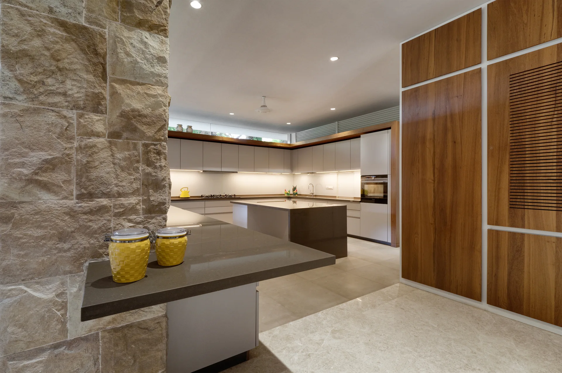A minimalist kitchen with a textured stone pillar, grey cabinets, a central island, and tall wood veneer built-ins under recessed lighting.