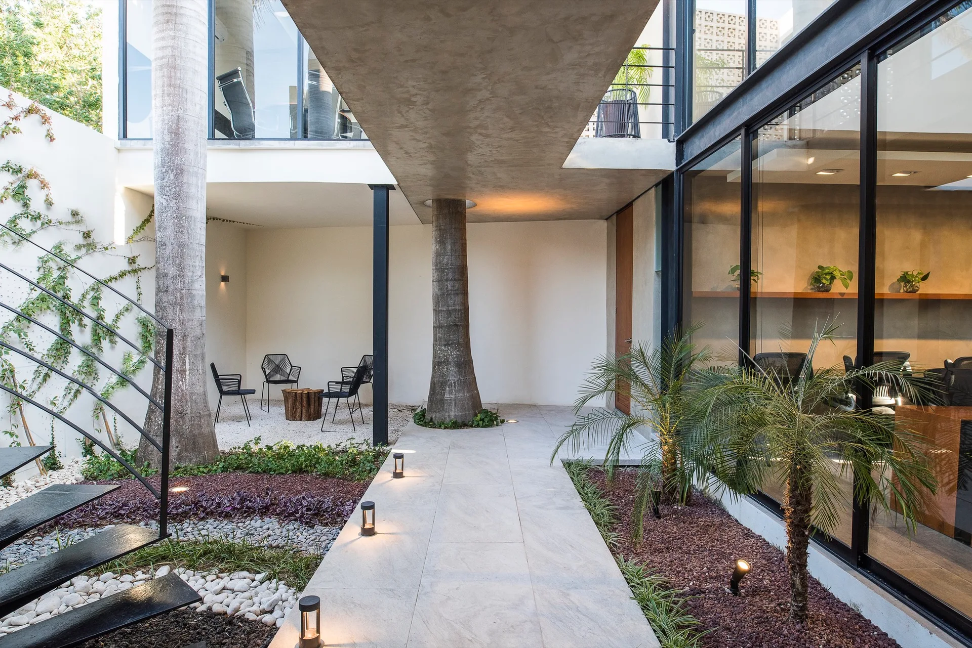 A modern paved walkway with low bollard lights leads through a landscaped courtyard under an exposed concrete canopy next to large glass walls.