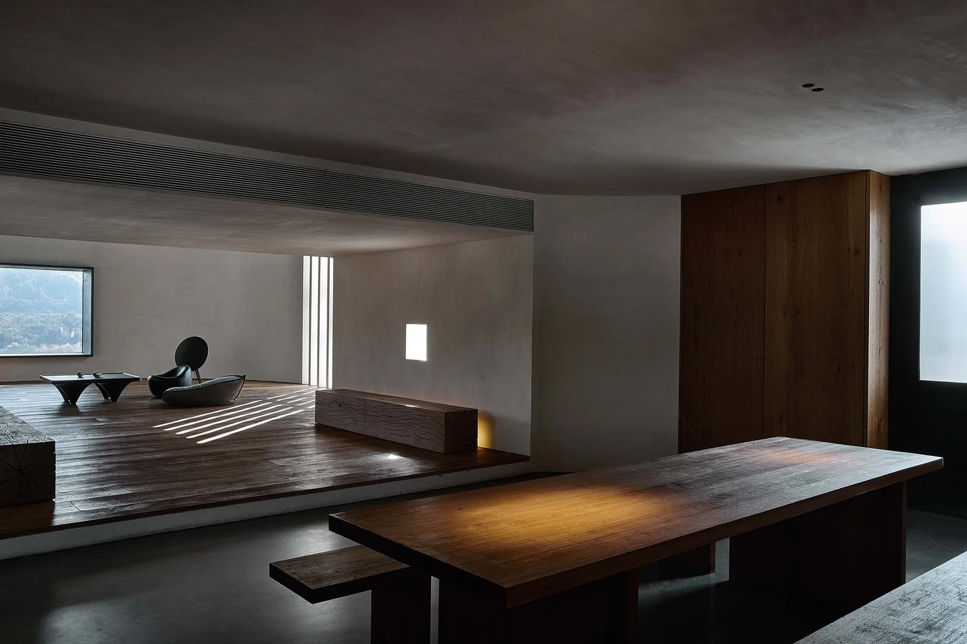 A minimalist interior featuring a dark wood dining table with benches and a raised wooden platform lounge area.
