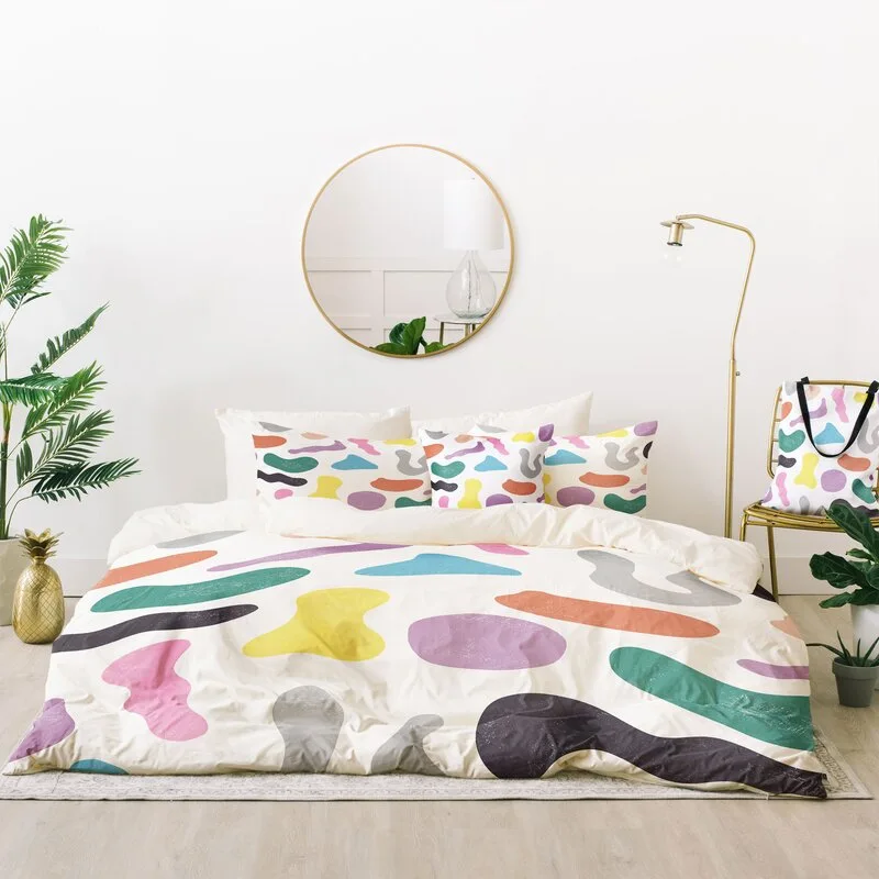 A bright bedroom features a bed with a multicolored abstract print duvet, a round gold-framed wall mirror, and a slim gold floor lamp.