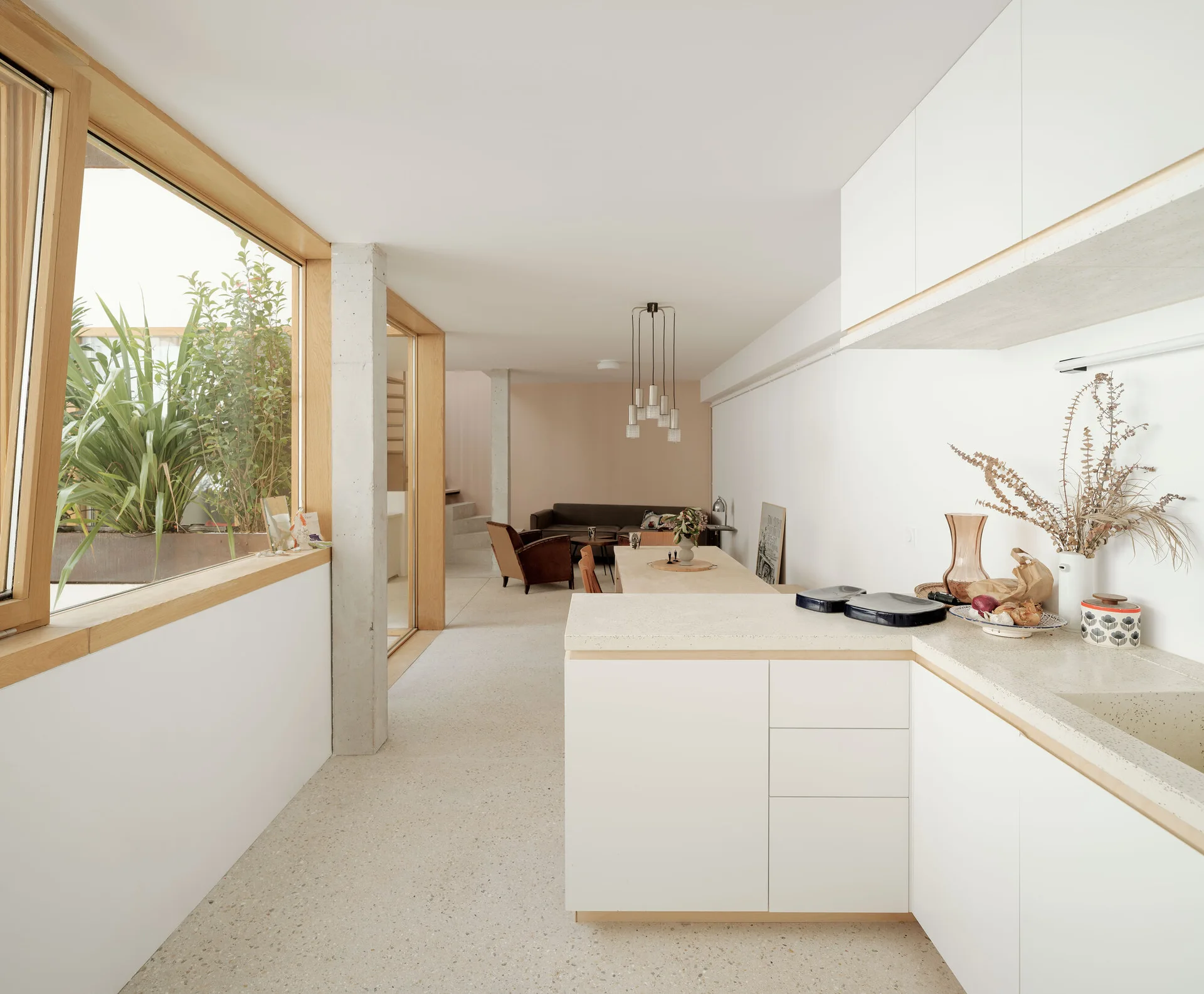 A minimalist white kitchen features terrazzo countertops, flat-front cabinets, and wood-framed windows overlooking a plant-filled courtyard.