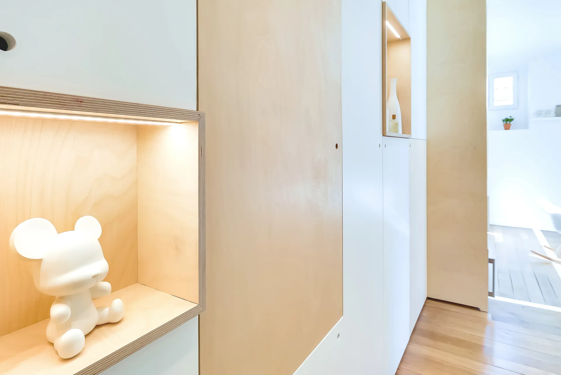 Close-up view of minimalist built-in cabinetry featuring white and light wood panels, exposed plywood edges, and warm-lit recessed display alcoves.