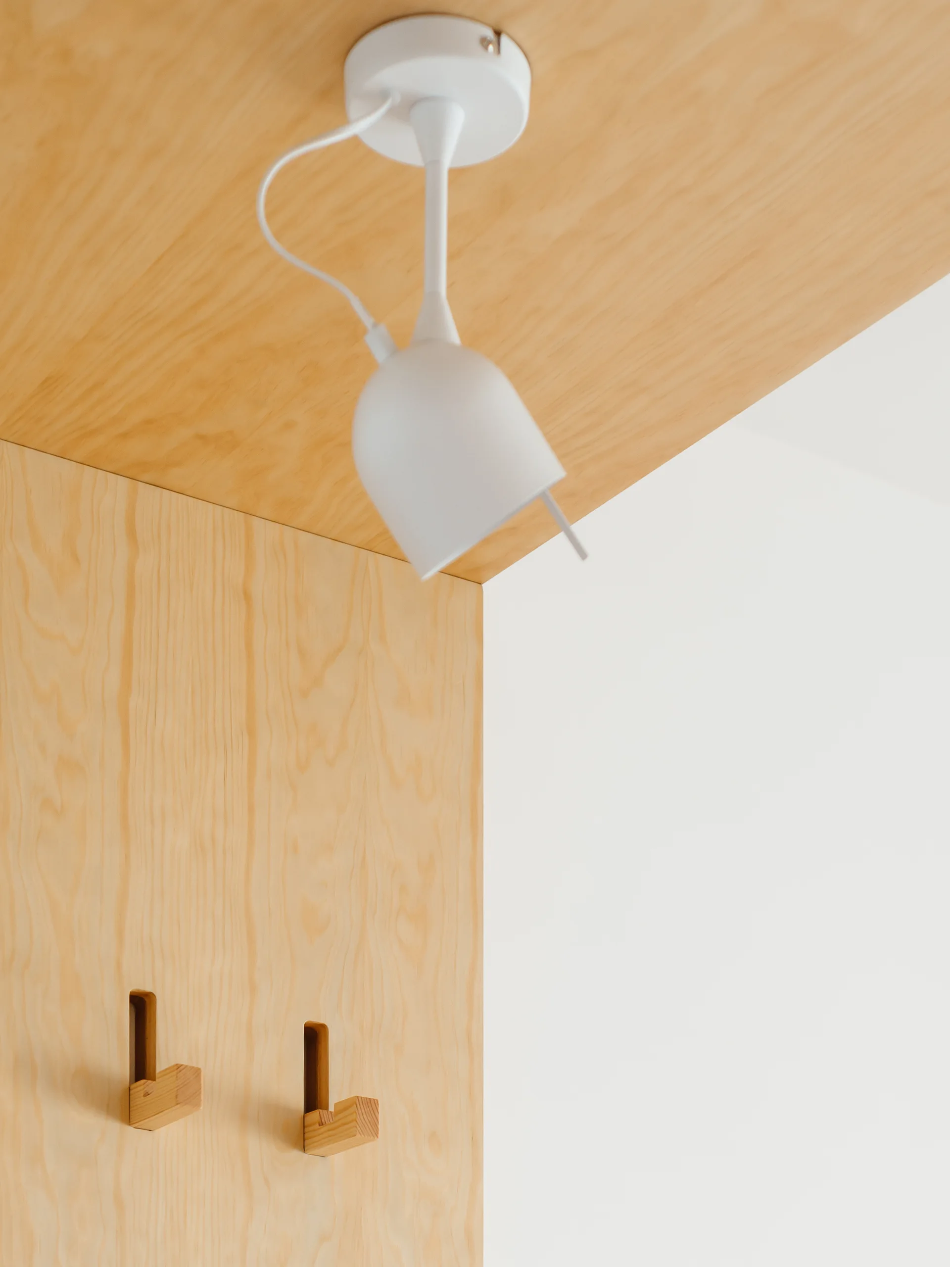 A white modern pendant light mounted on a light plywood ceiling next to a matching plywood wall with integrated wooden coat hooks.