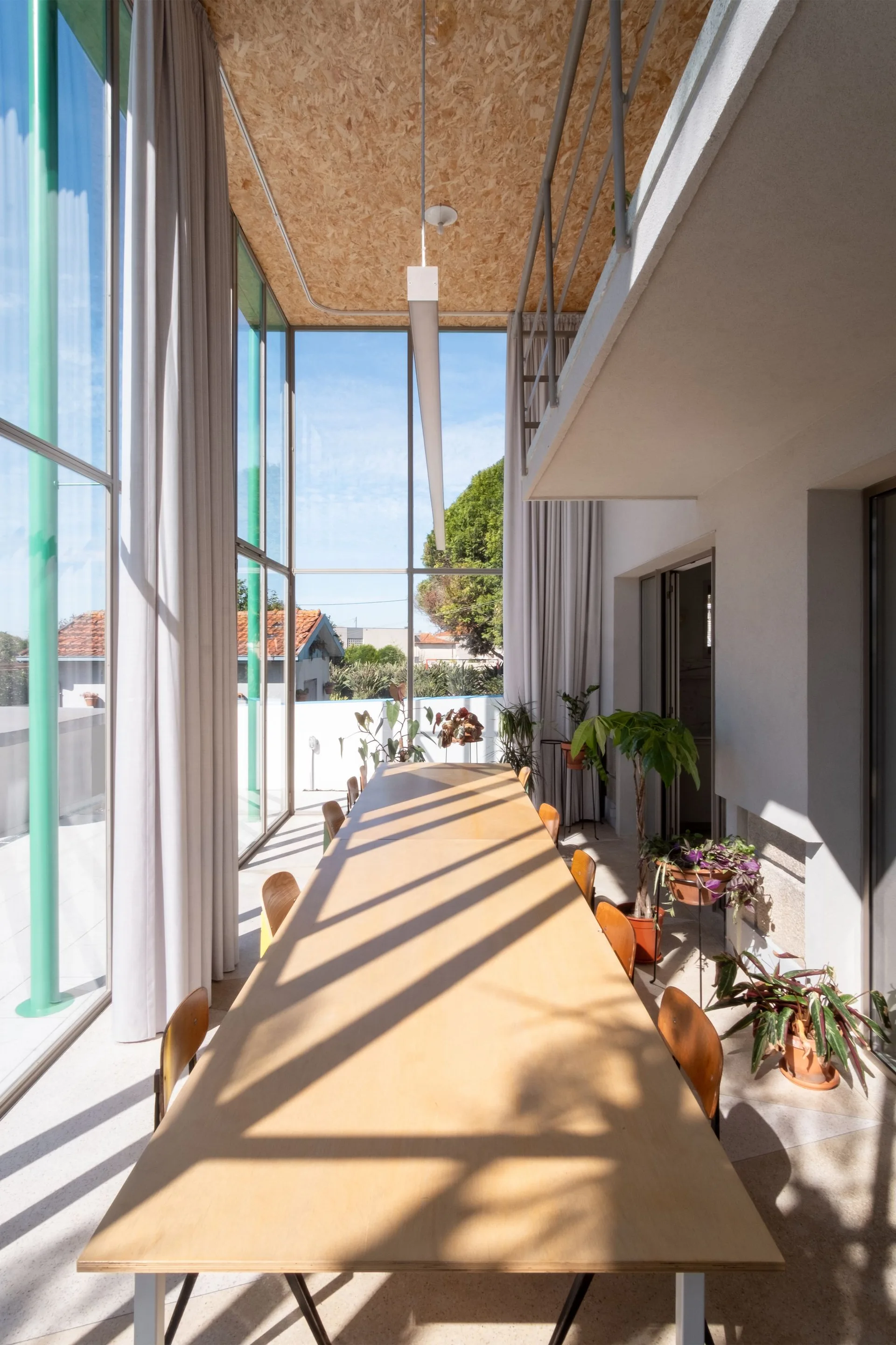 A long wooden dining table with chairs sits in a double-height dining room featuring floor-to-ceiling windows and an OSB ceiling.