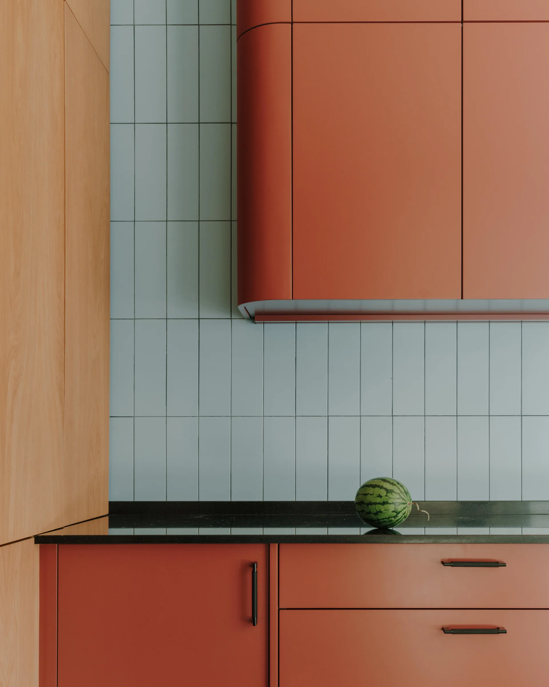 A modern kitchen details matte rust orange cabinets, a black countertop, and a vertically stacked light blue subway tile backsplash.