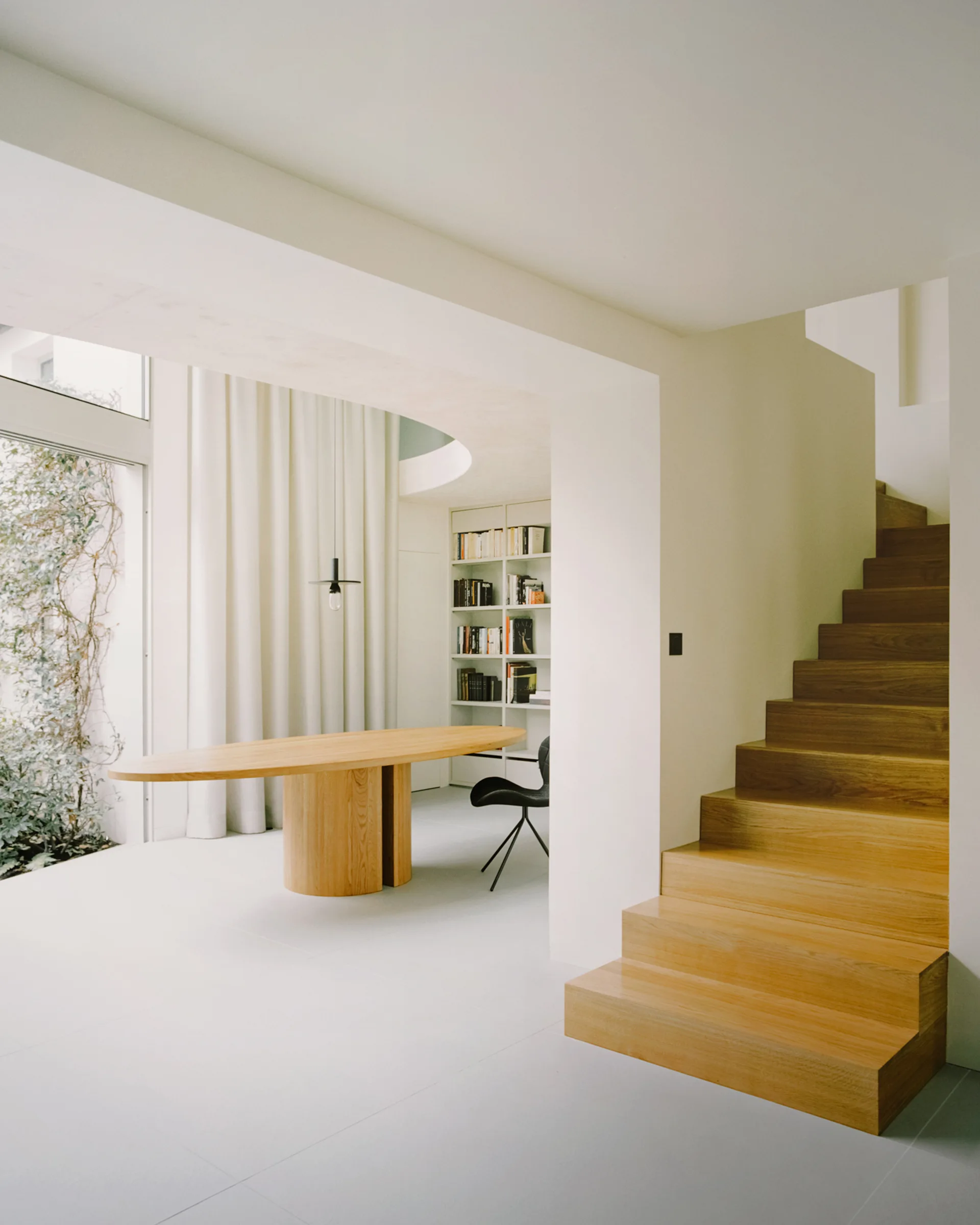 A bright white minimalist dining space features an oval wooden table next to a prominent wooden staircase and a recessed bookshelf.
