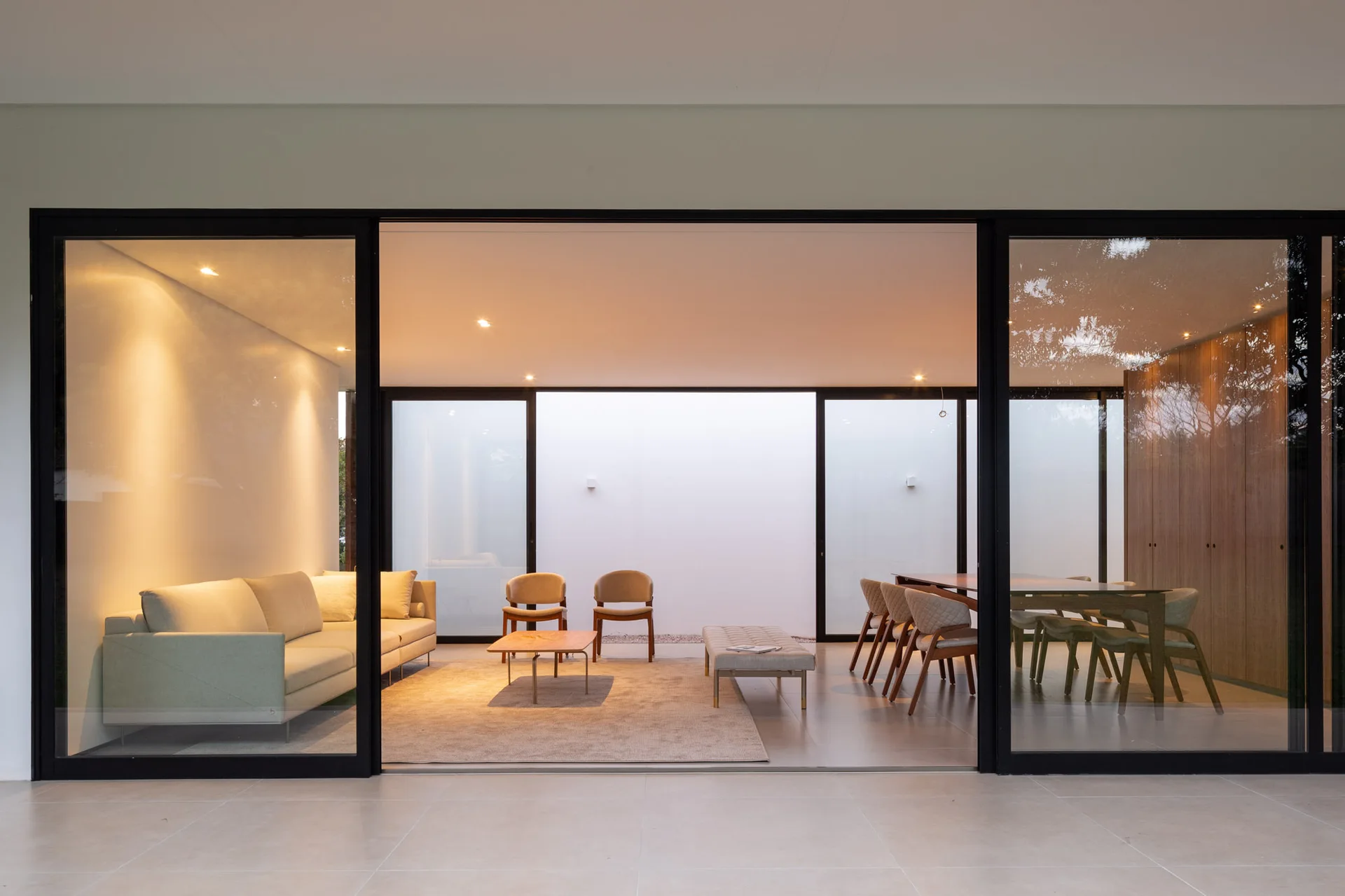 A modern open-concept living and dining area with warm lighting viewed through large black-framed sliding glass doors.