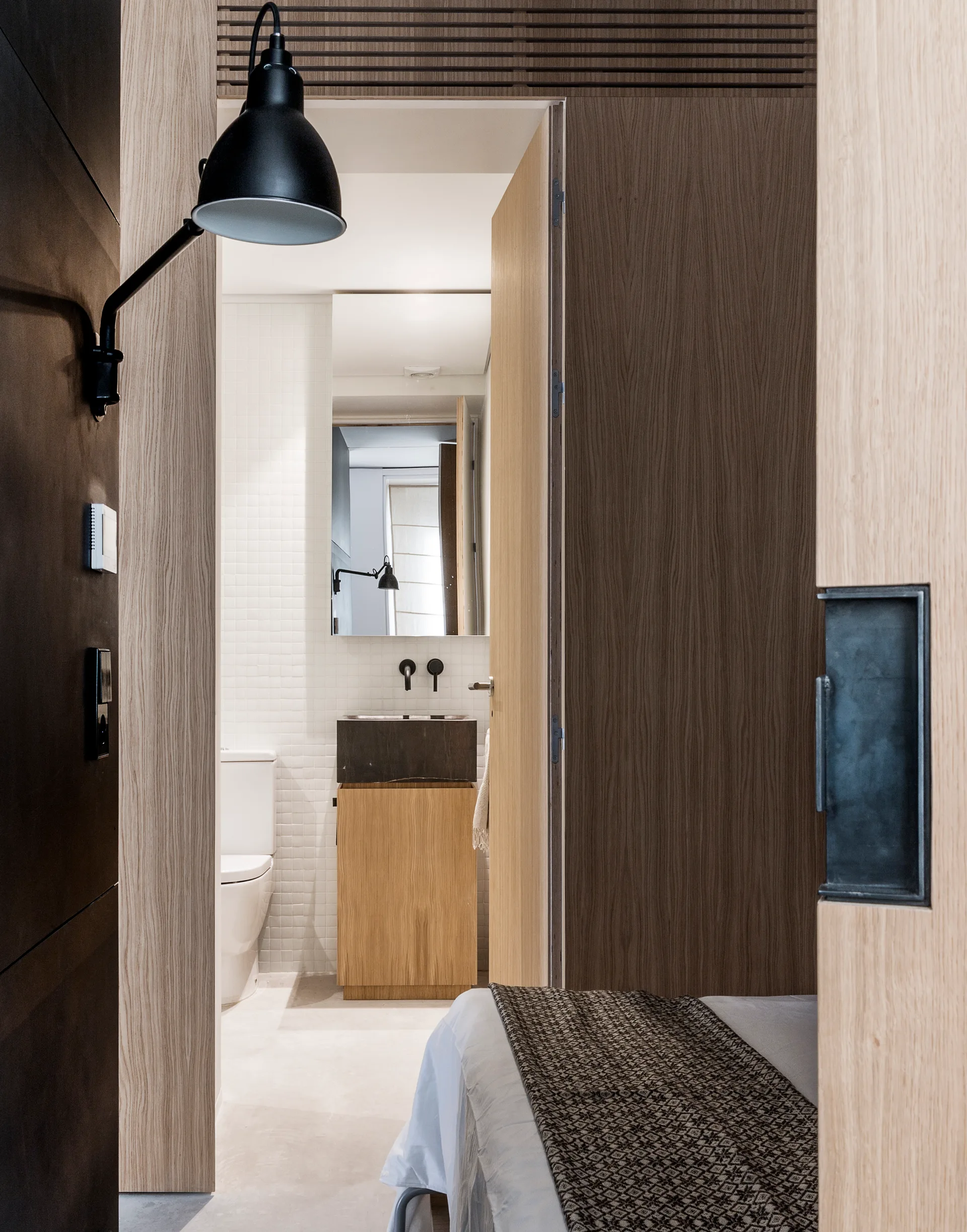 A view through a wood-paneled doorway into a bathroom with white grid tile walls, a wood vanity, and a black block sink.