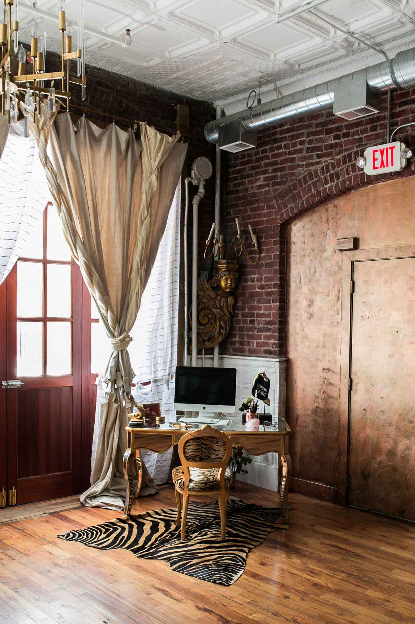 A home office space in an industrial loft features a gold desk, a tiger-print chair and rug, exposed brick walls, and a pressed tin ceiling.