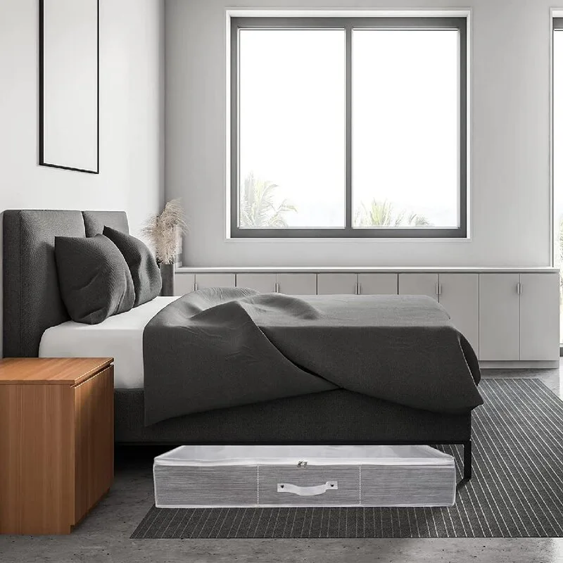 A grey upholstered bed with a dark grey duvet features a grey zippered fabric storage bin tucked underneath on a striped rug.