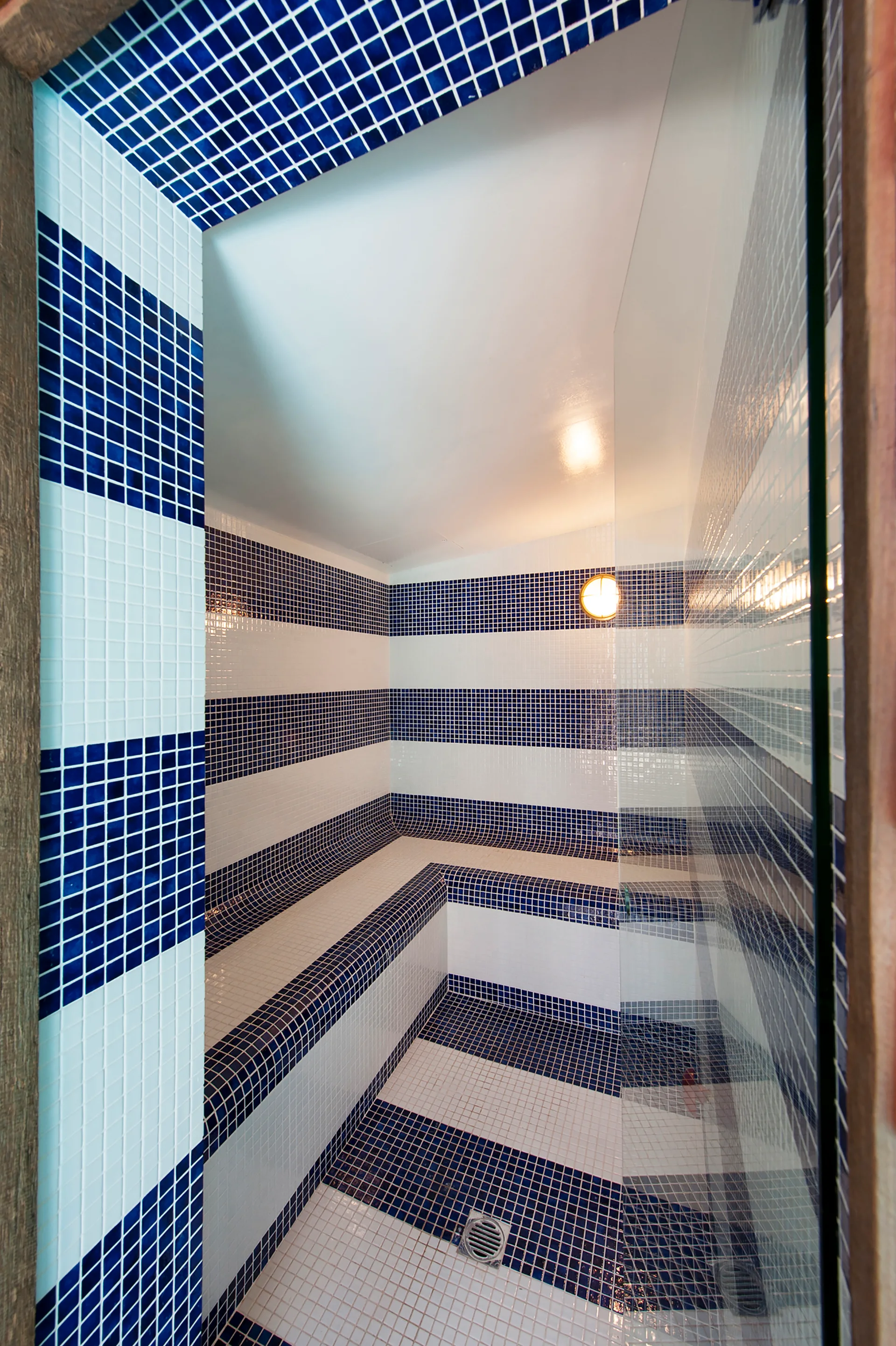 A steam room features horizontal blue and white stripes made of small mosaic tiles covering the walls, floor, and built-in bench.