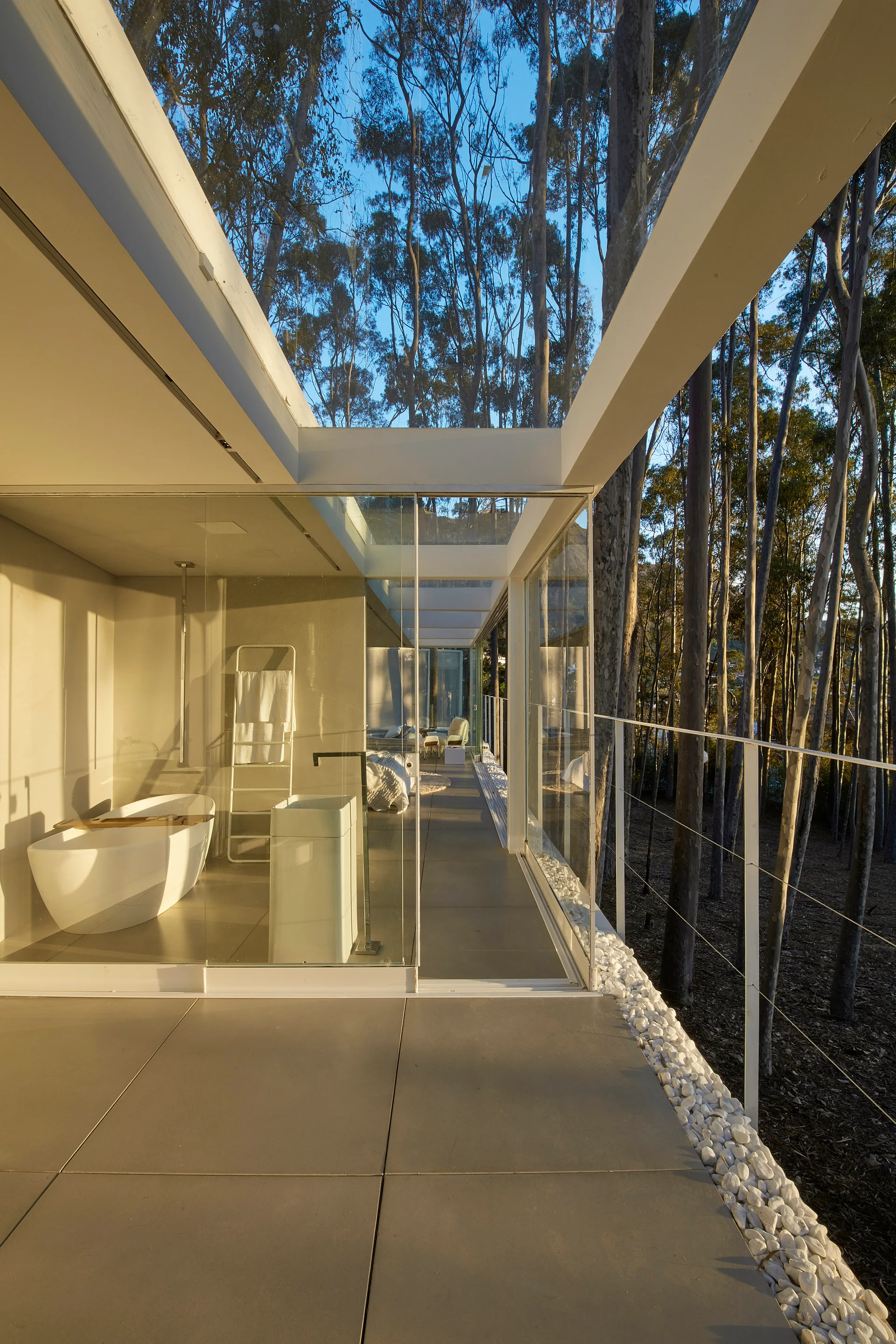 A minimalist bathroom with a freestanding tub and glass walls looking out onto a modern balcony with white pebbles and surrounding trees.