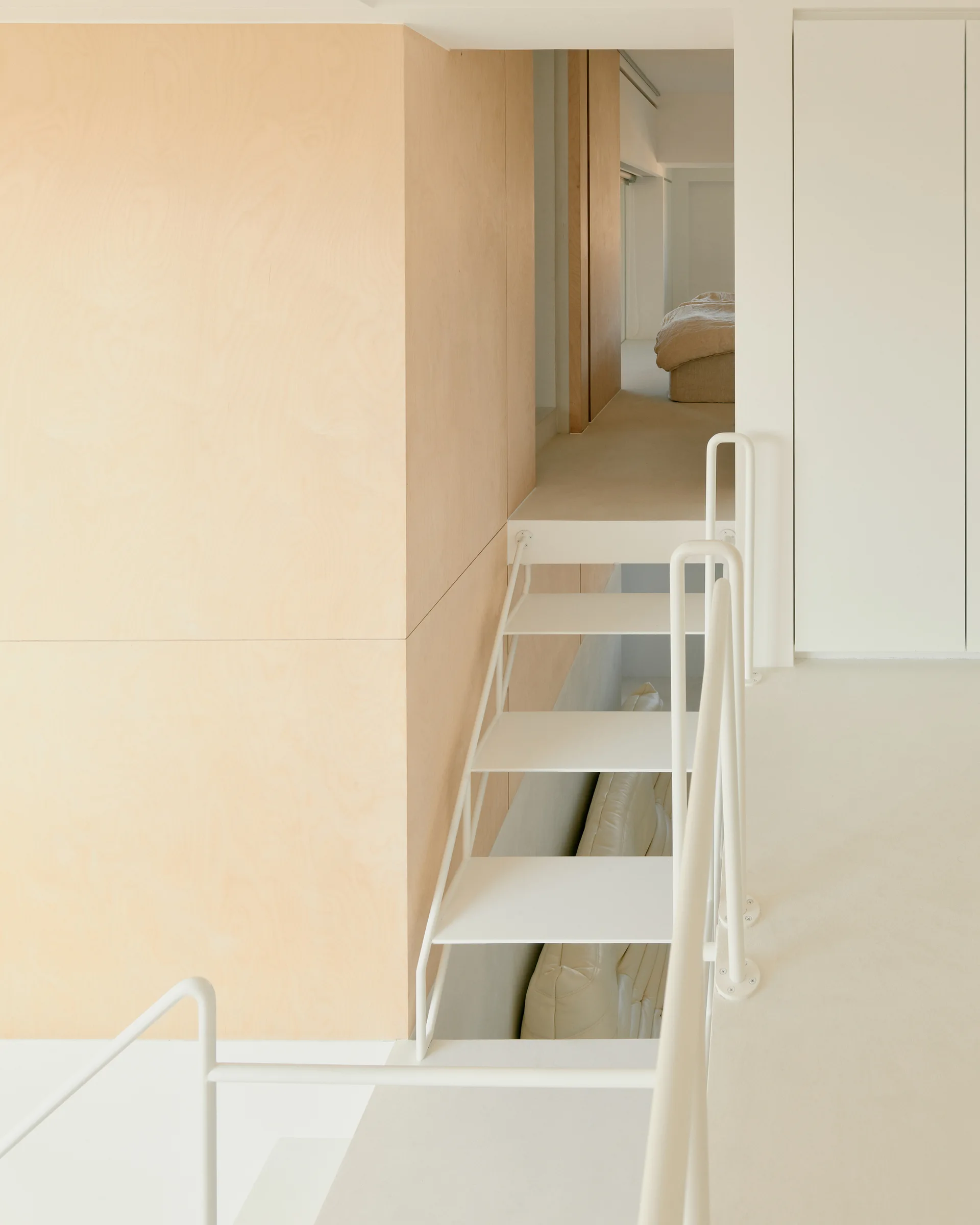 An open-tread white metal staircase with simple railings sits next to a light plywood wall in a minimalist split-level interior.