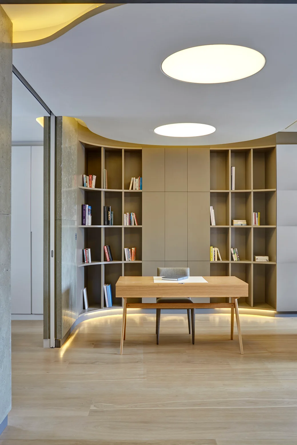 A minimalist home office features a wooden desk centered in front of a curved tan built-in bookcase wall illuminated by recessed lighting.