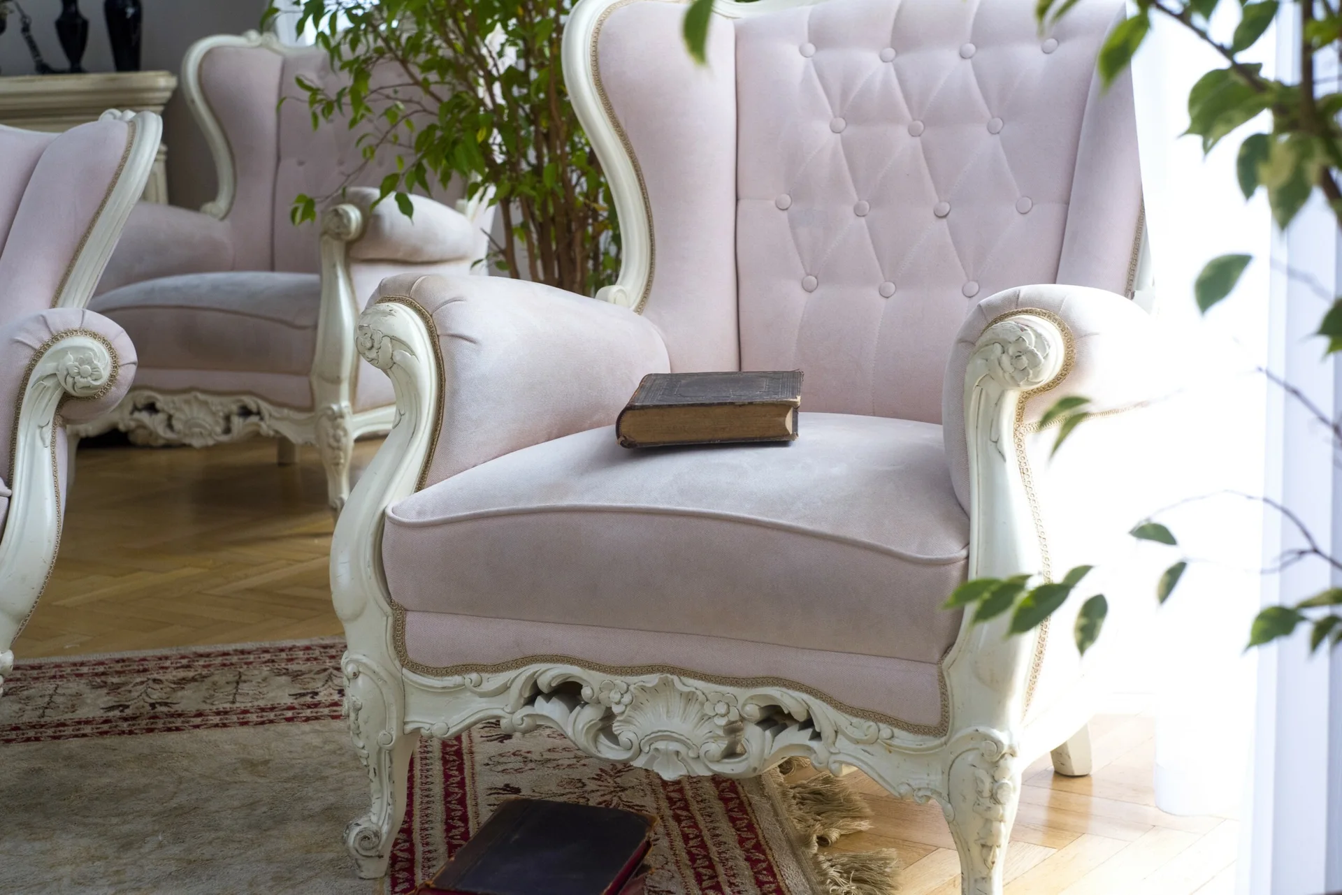 Two light pink tufted armchairs with white ornate carved wood frames sit on a patterned area rug over hardwood flooring.