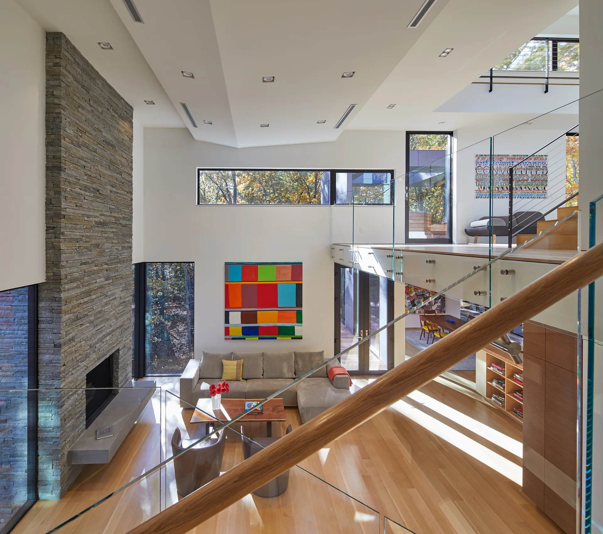 A double-height modern living room featuring a tall stacked-stone fireplace, a grey sectional sofa, and a mezzanine balcony with glass railings.