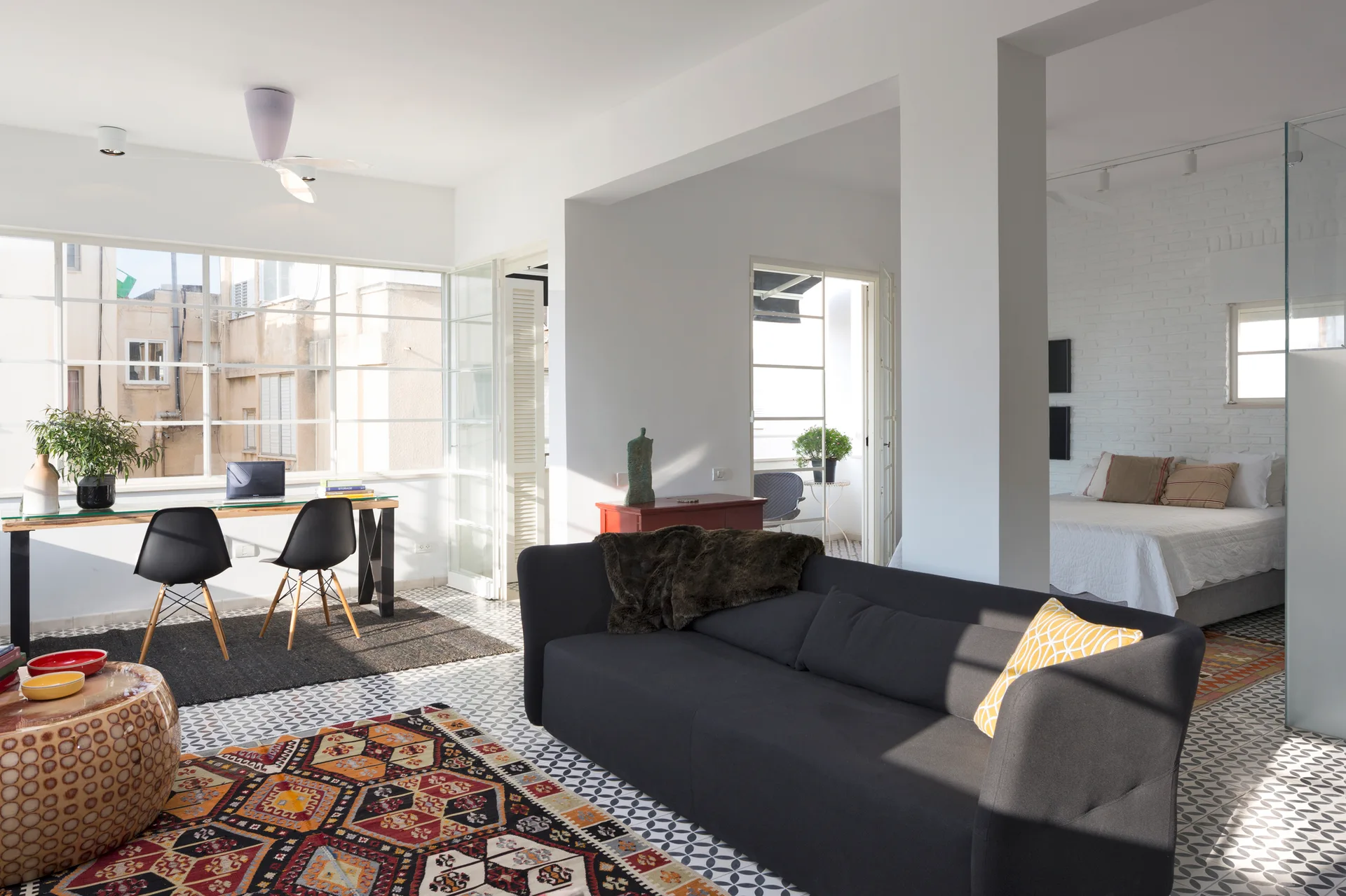 A bright open-plan living room featuring a dark sofa, geometric tile flooring, a glass-top office desk, and a bedroom visible in the background.