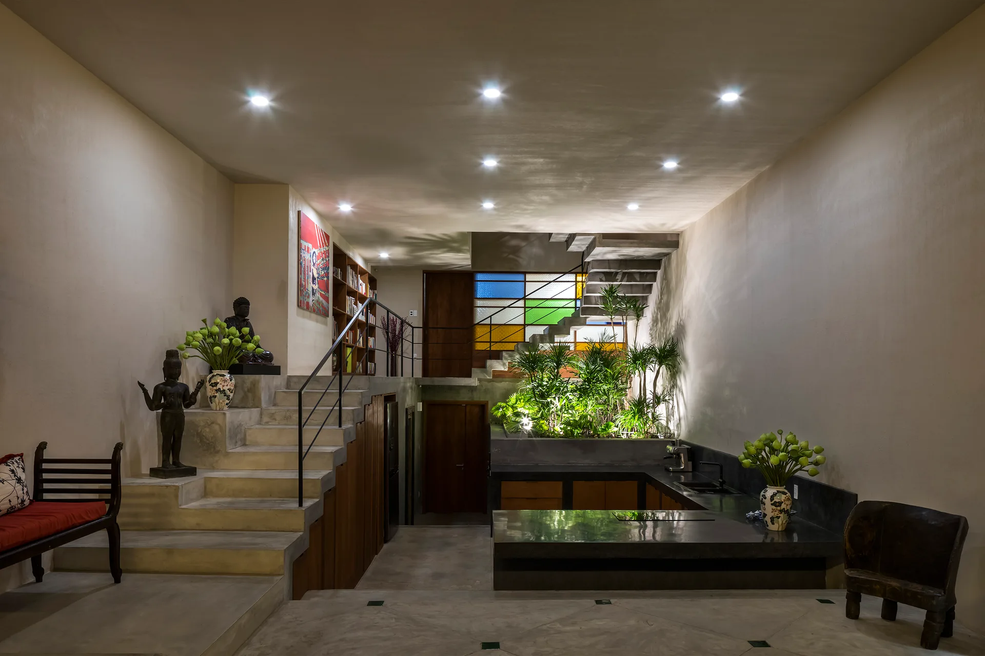 A split-level concrete interior features a staircase leading to a mezzanine, a dark kitchen counter, and an illuminated indoor planter with green foliage.