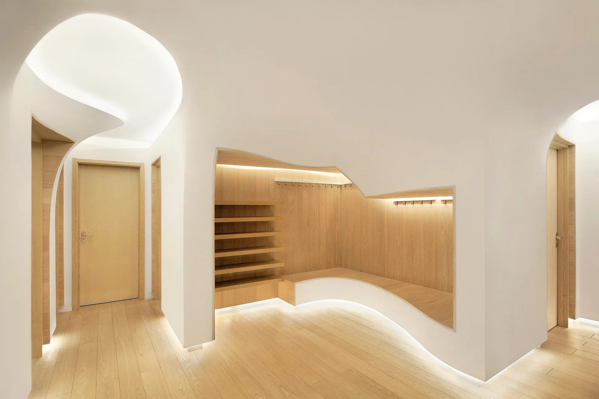 A minimalist mudroom features curved white plaster walls, a light oak built-in storage bench, open shelves, and glowing LED strip lighting.