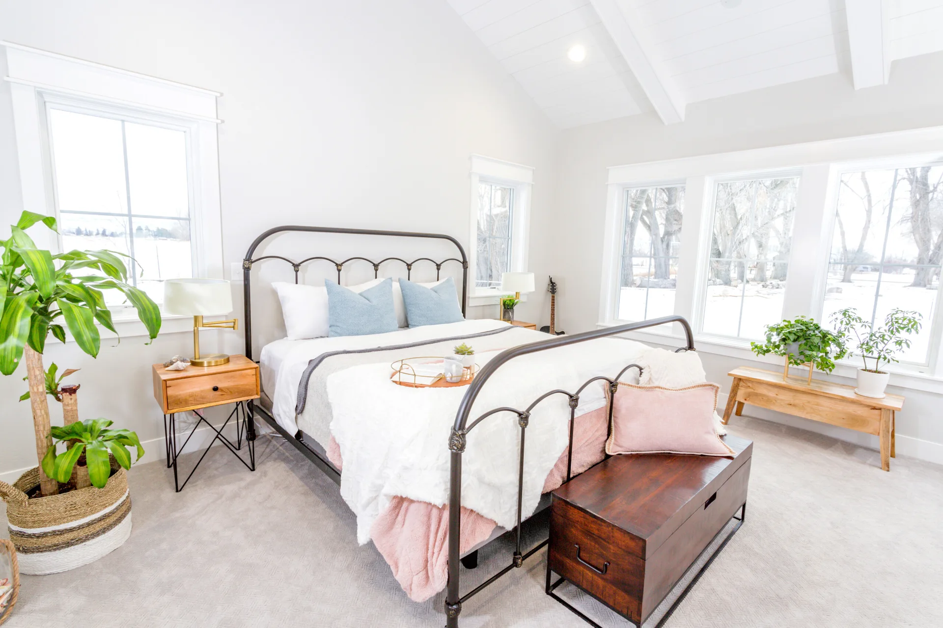 A bright bedroom features a black metal bed frame, pastel bedding, a dark wood storage chest, and a vaulted ceiling with an exposed beam.