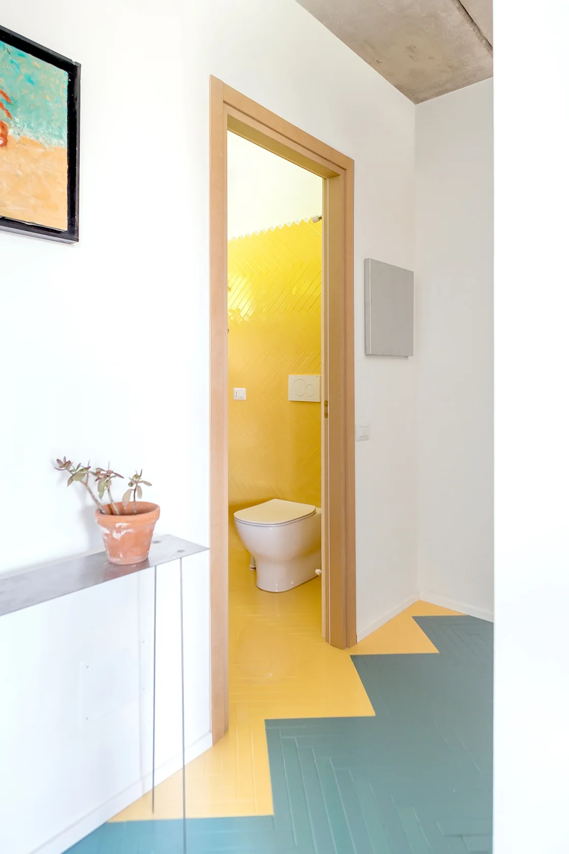 A view through a wooden doorway showing a yellow tiled bathroom and a hallway floor with transitioning teal and yellow herringbone tiles.