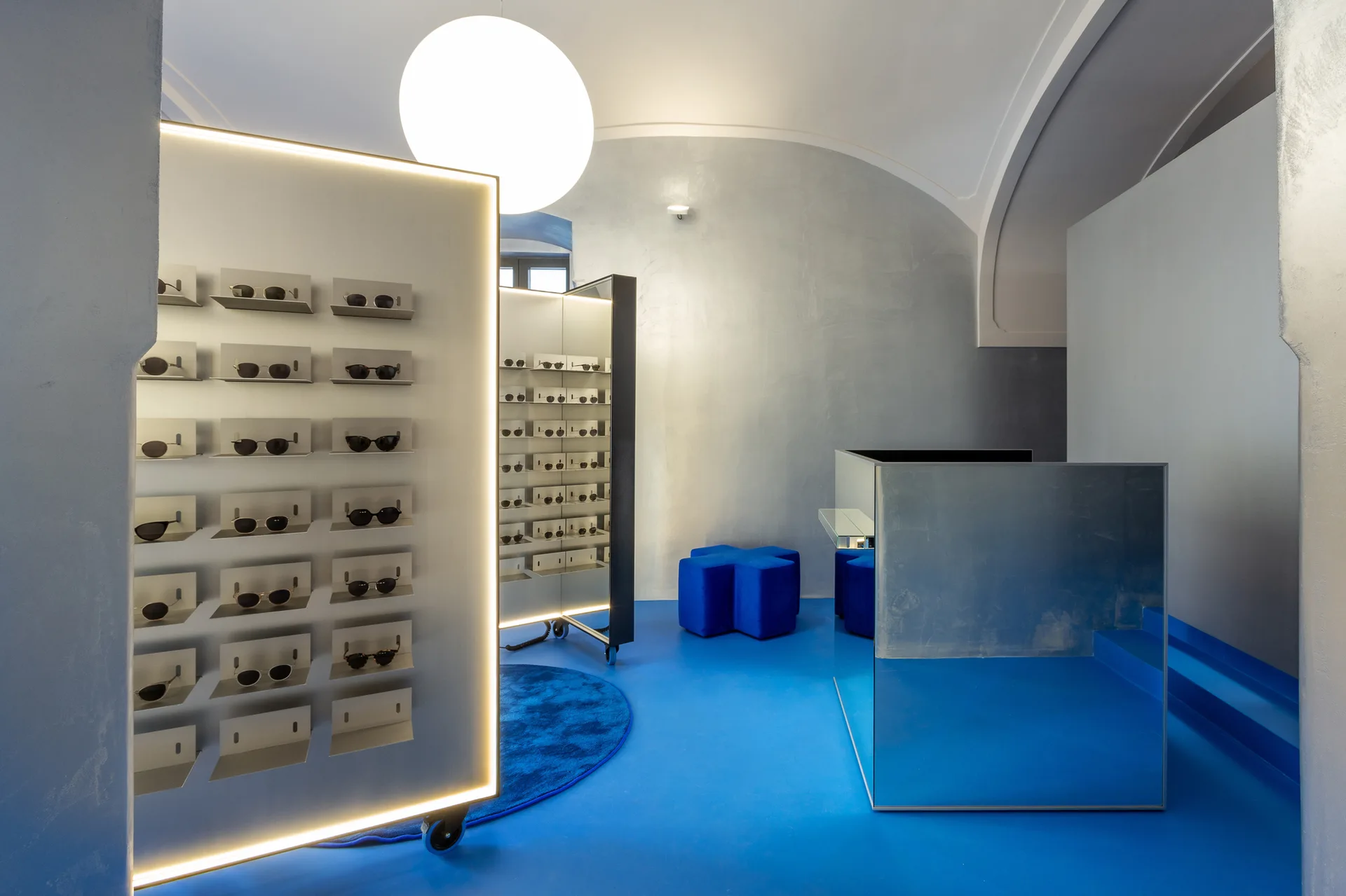 A minimalist retail showroom features a vibrant blue floor, rolling illuminated sunglass displays, and a reflective mirrored counter.