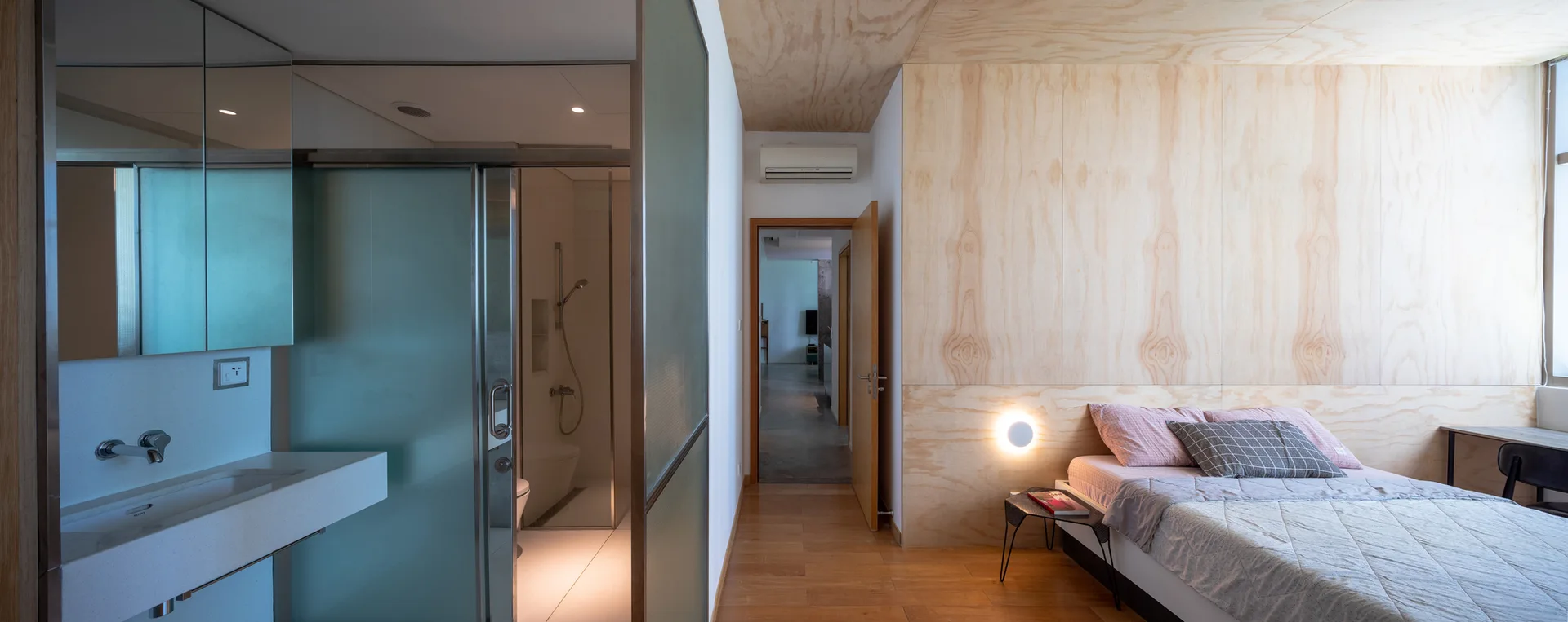 A minimalist bedroom featuring plywood wall panels, a bed with grey bedding, and an adjacent bathroom with a frosted glass sliding door.