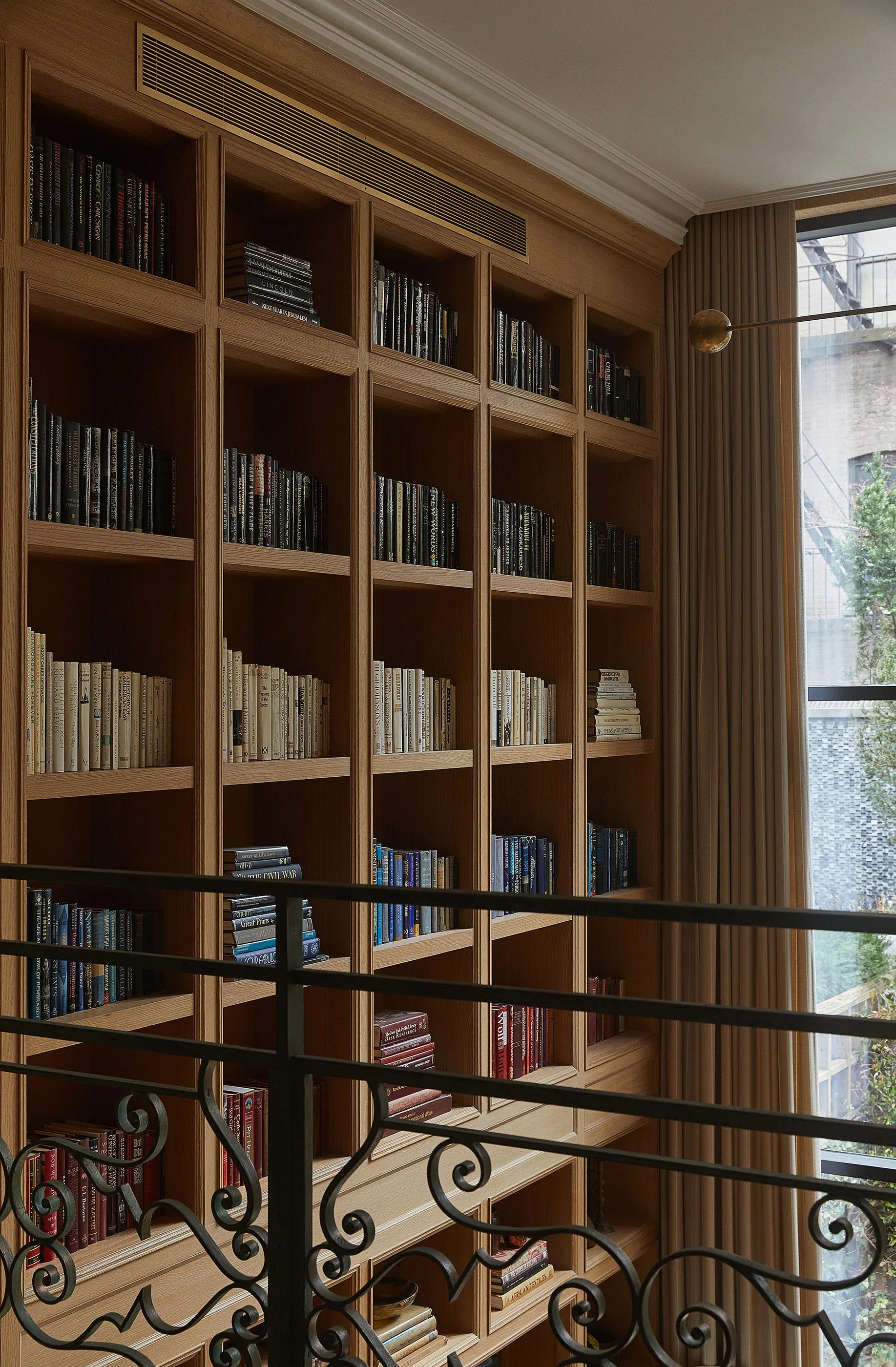 Floor-to-ceiling wooden bookshelves filled with books stand next to a draped window behind a dark wrought iron railing.
