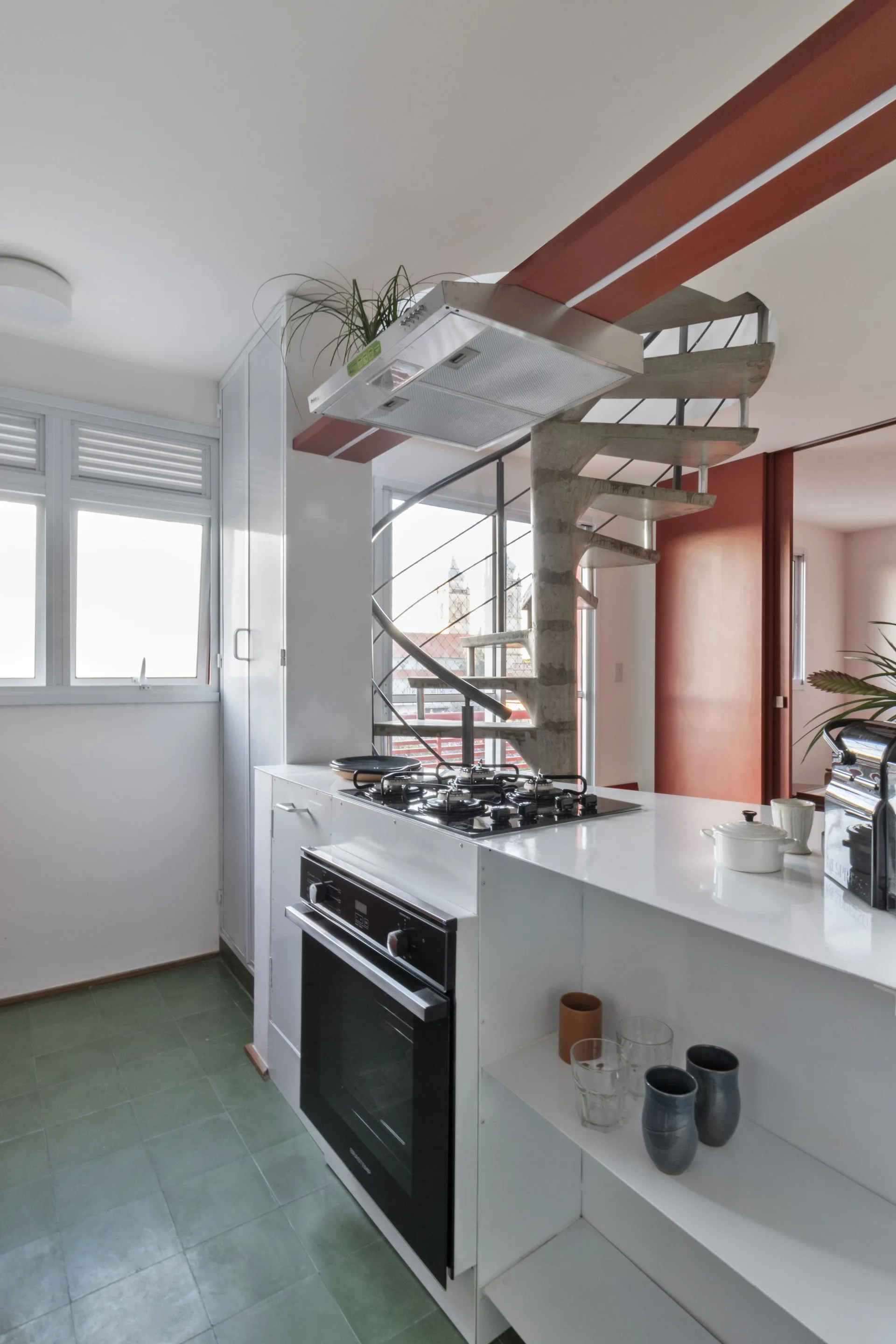 A modern white kitchen features green floor tiles, a white island with a cooktop, and a concrete spiral staircase in the background.