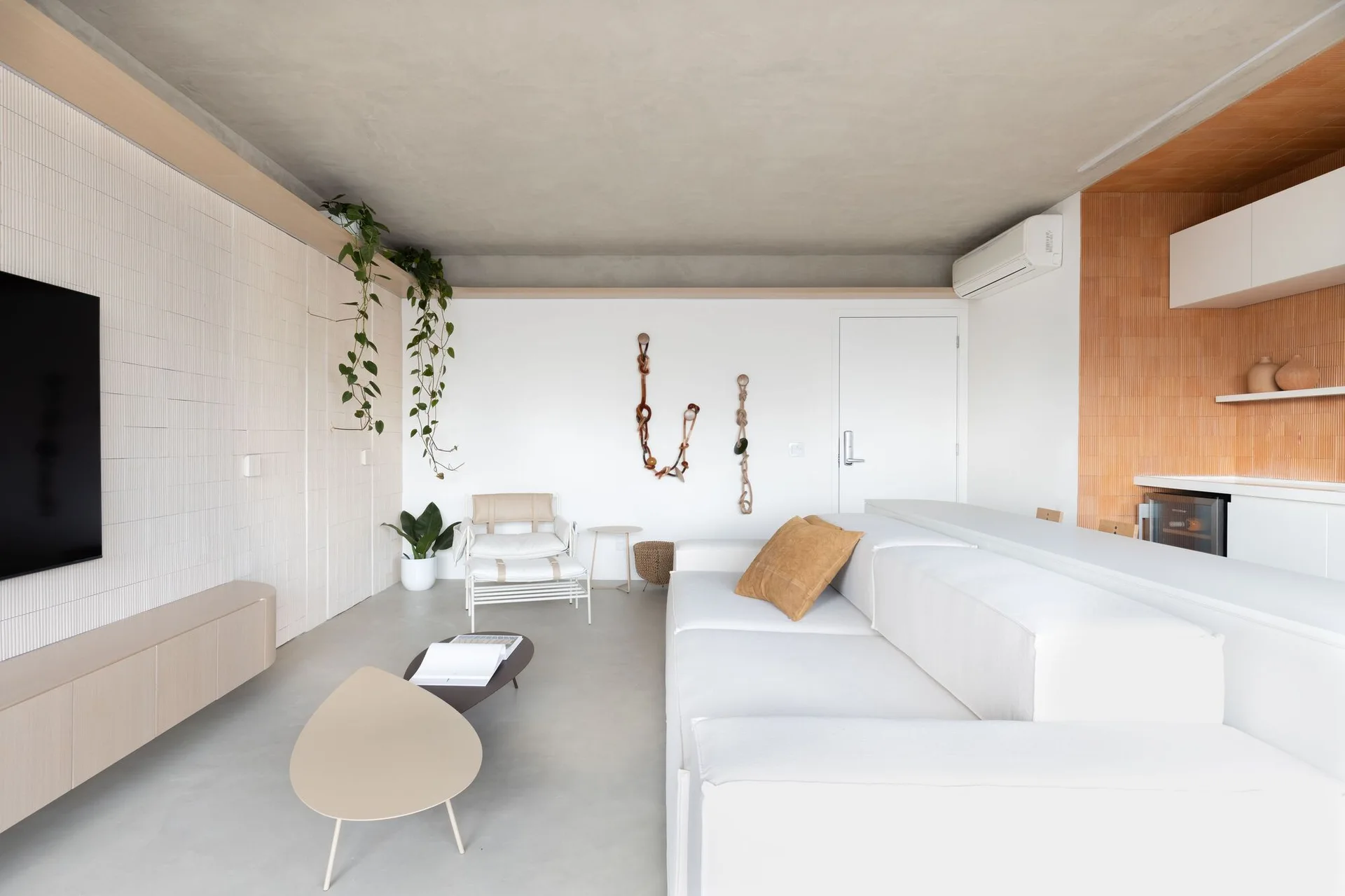 A minimalist living room features a white sectional sofa, a smooth concrete ceiling, and vertical slatted wall paneling with hanging green plants.