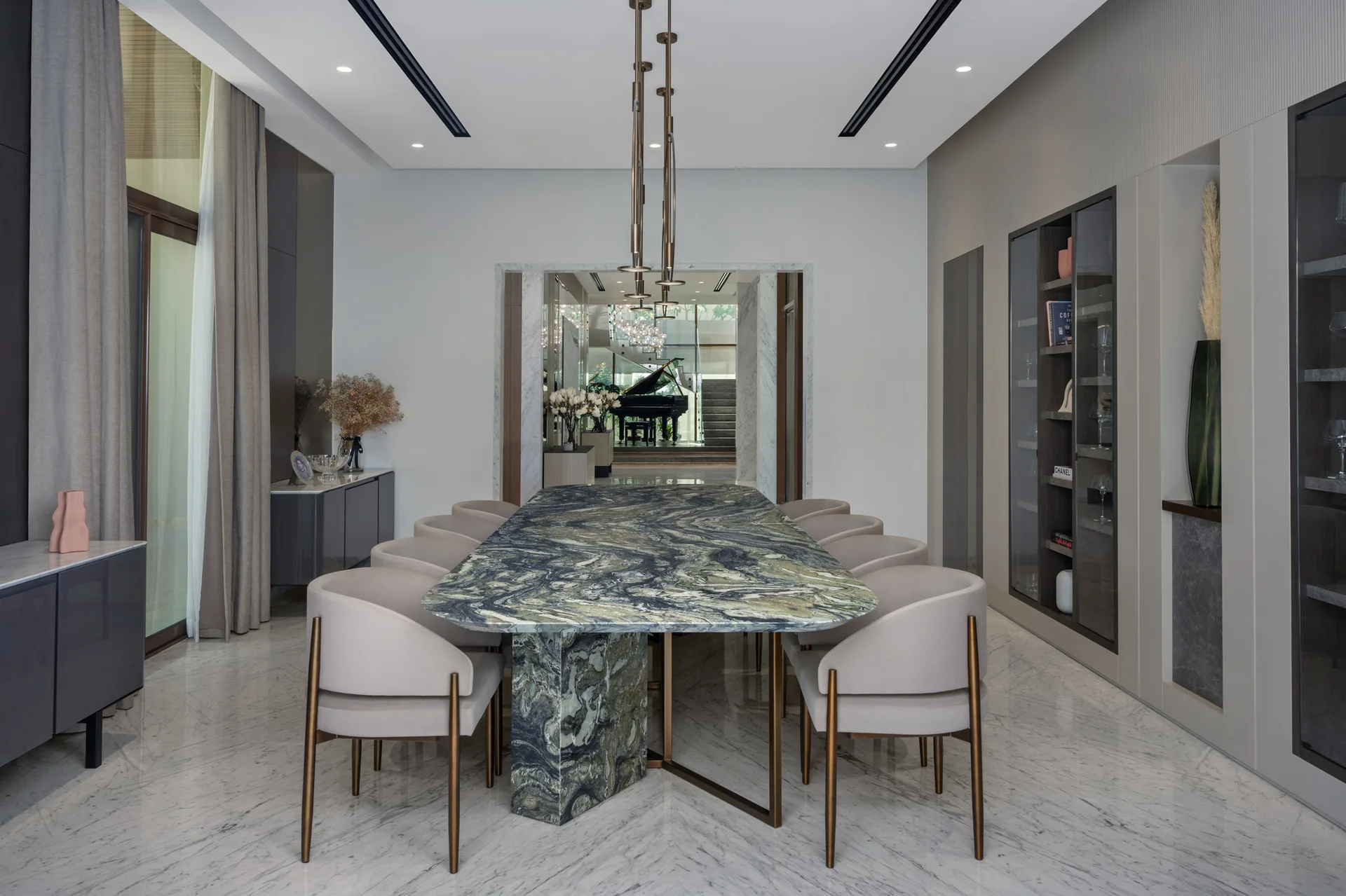 A spacious dining room featuring a green marble dining table with beige chairs, polished white stone flooring, and integrated wall shelving.