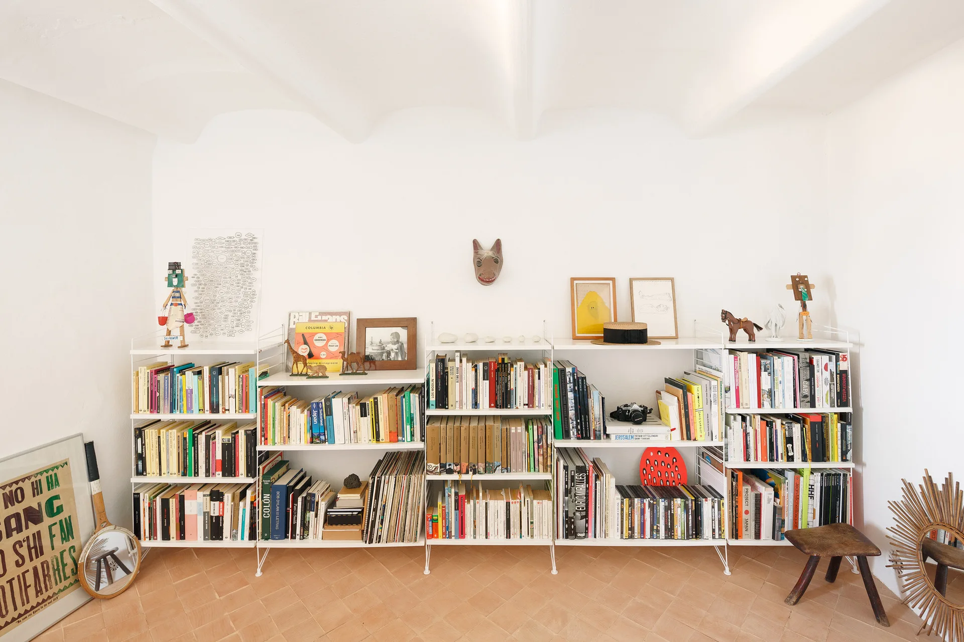 A white modular wire shelving unit holding books, vinyl records, and art pieces stands against a white wall on a terracotta tiled floor.