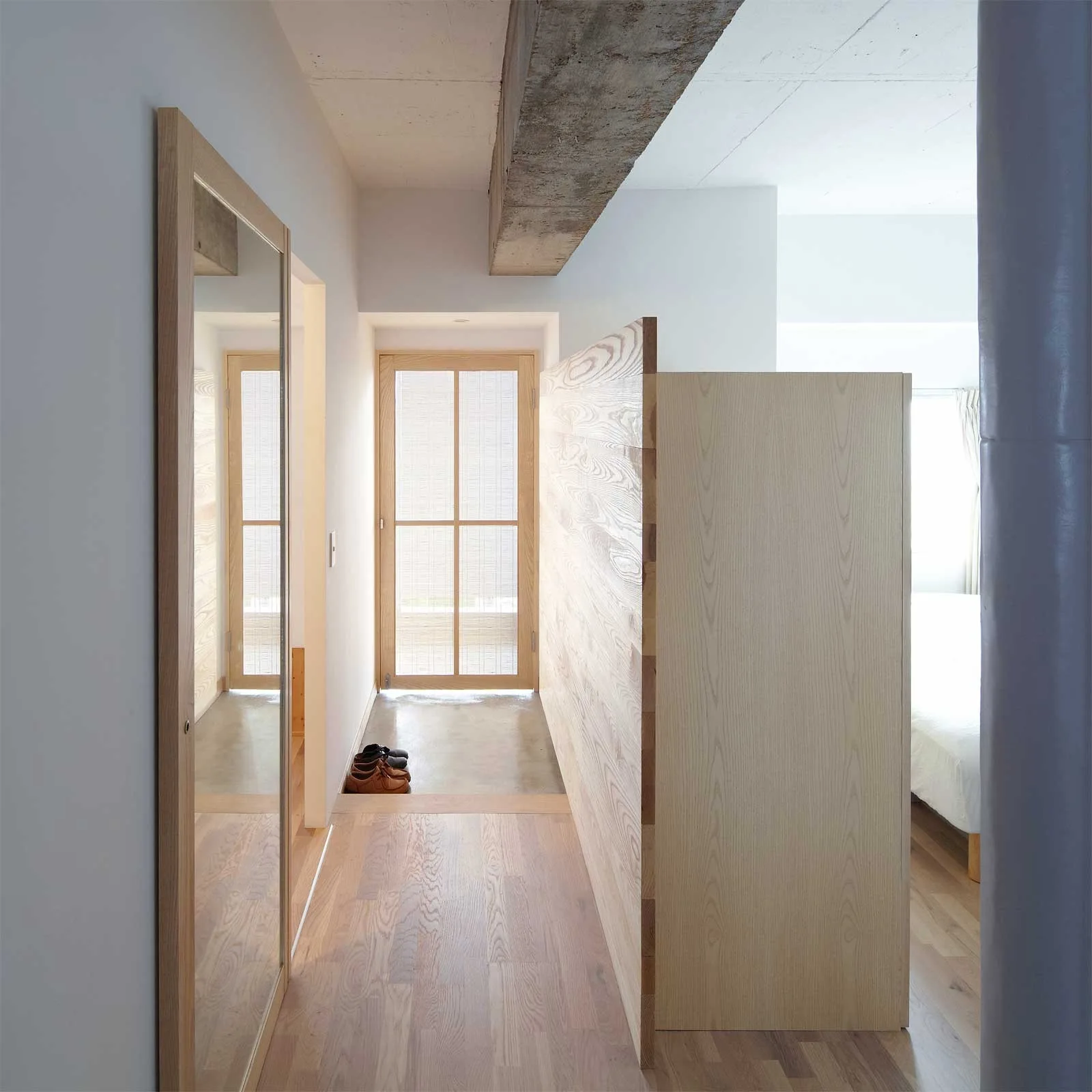 A minimalist entryway with light wood flooring, a tall wooden partition wall, a wall mirror, and an exposed concrete ceiling beam.