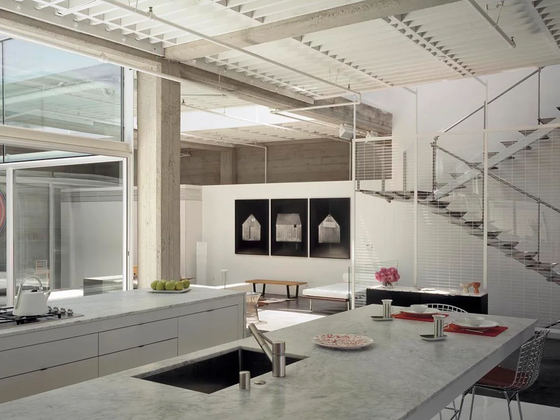 An industrial loft kitchen featuring two large white marble islands, an exposed concrete column, and a minimalist steel staircase in an open floor plan.
