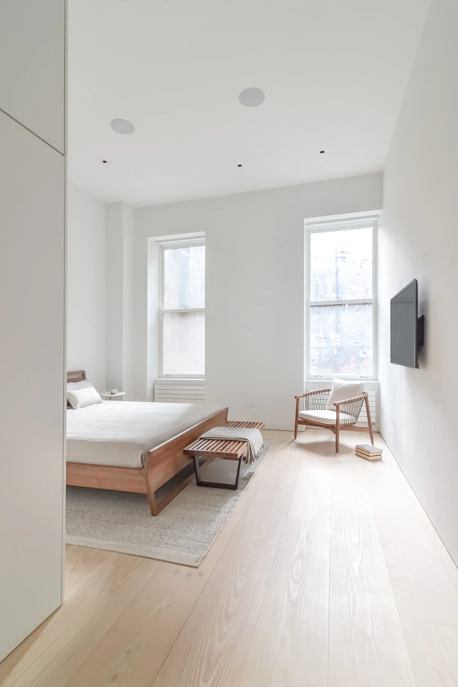 A minimalist bedroom features a wooden bed frame with a slatted bench, a woven armchair near two tall windows, and light blonde wood flooring.