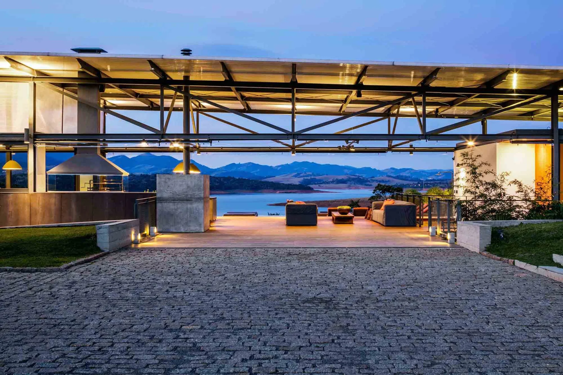 A modern open-air patio with a steel truss ceiling, wood decking, and grey outdoor sofas overlooks a quiet lake and mountains at dusk.