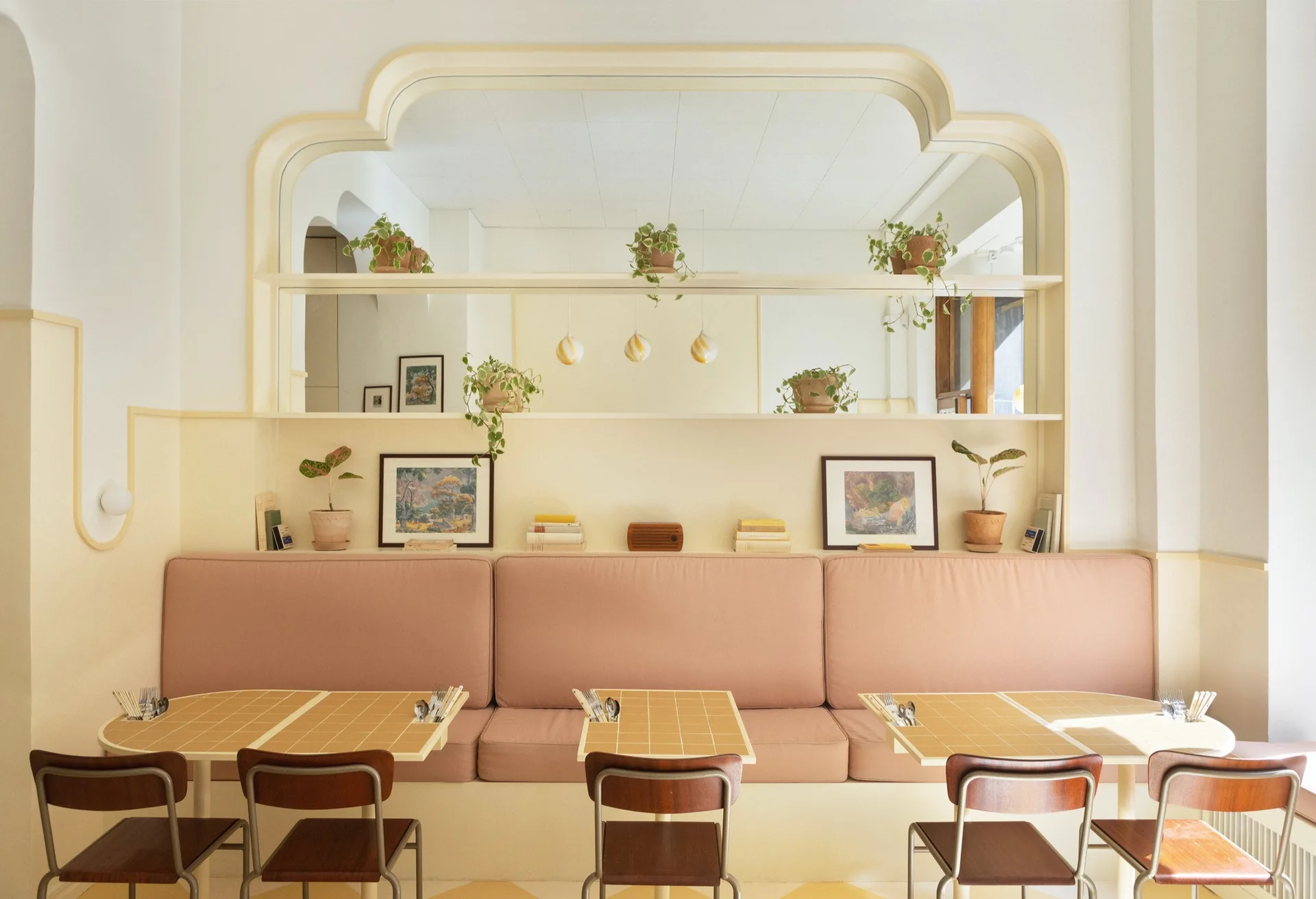 A pastel pink banquette sits against a cream wall with an arched mirror and a display shelf holding potted plants above tiled dining tables.
