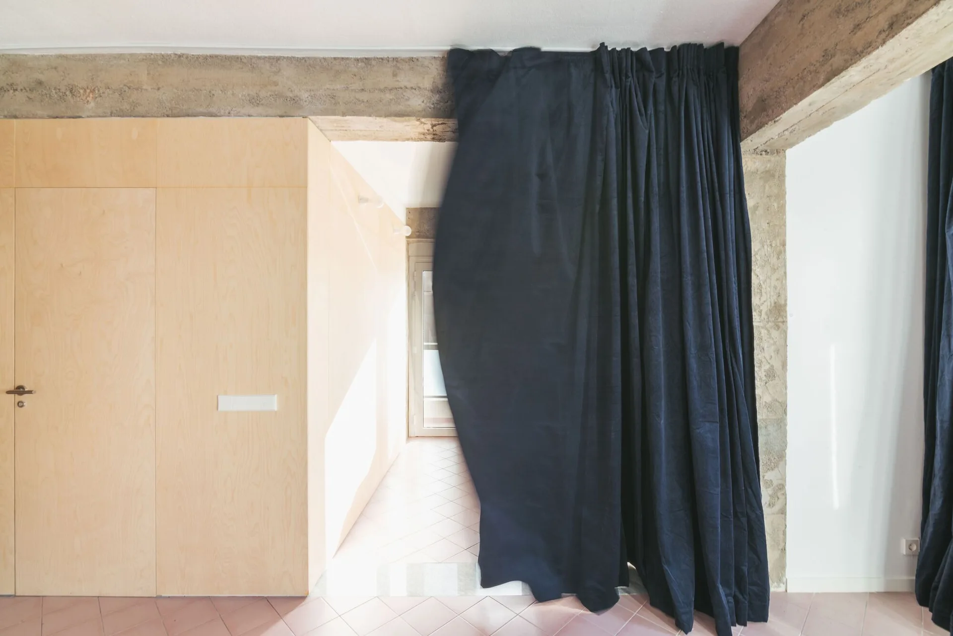 A transition space features light wood wall paneling, an integrated flush door, and a heavy navy blue fabric curtain draped below an exposed concrete beam.
