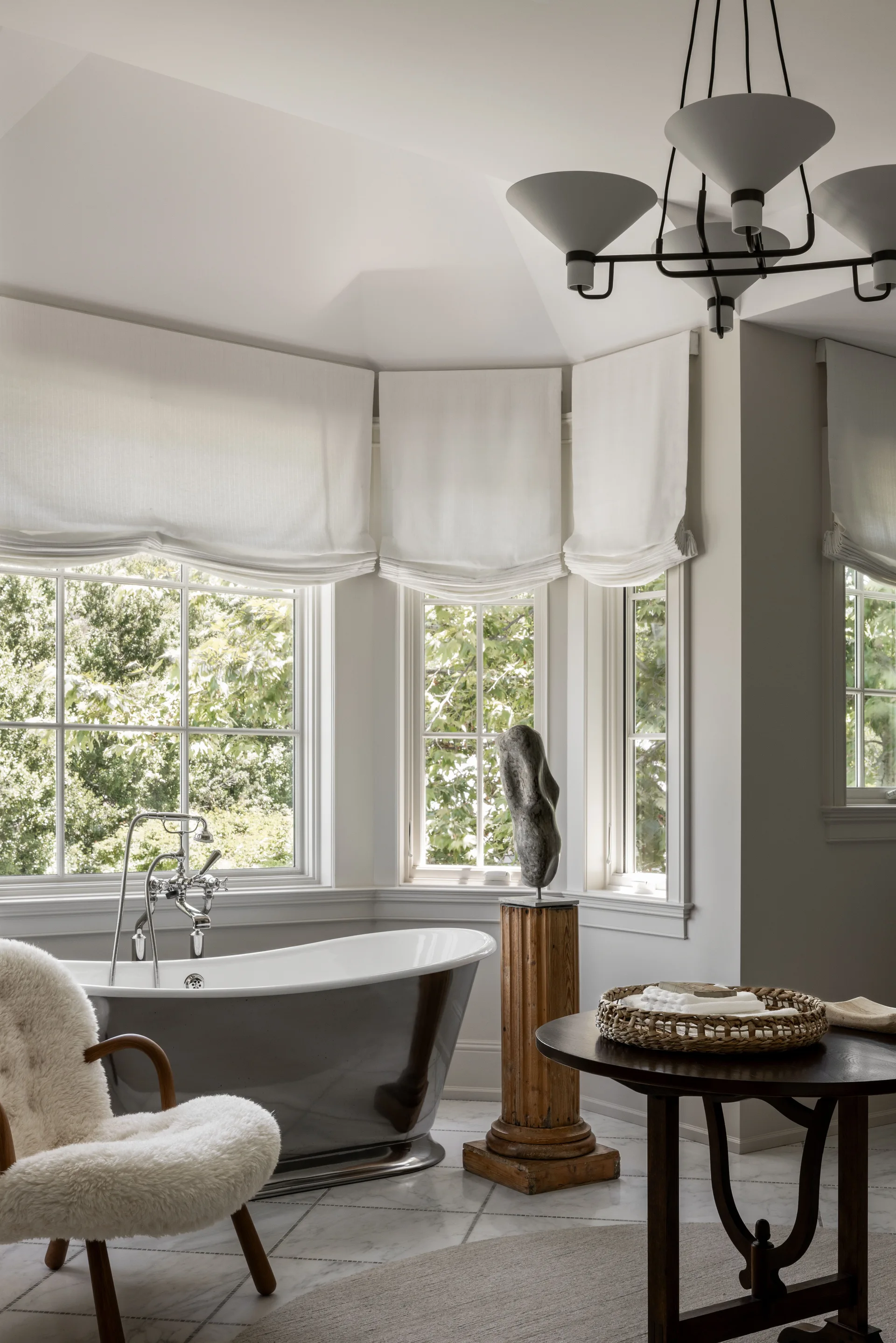 A dark grey freestanding bathtub sits in a bright bay window alcove alongside a wooden pedestal sculpture, a fluffy armchair, and a side table.