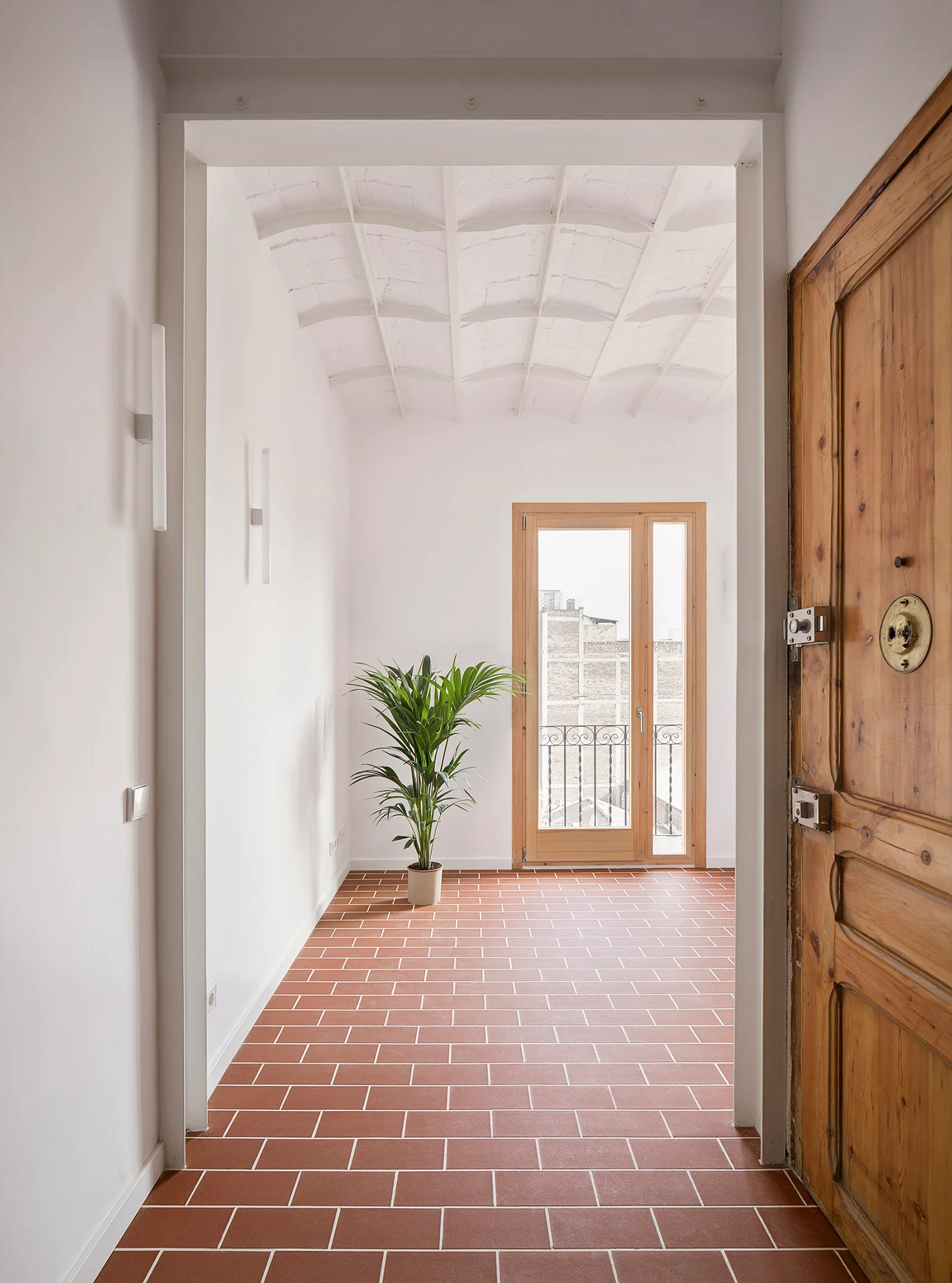 An entryway featuring a terracotta tile floor, a rustic wooden front door, white vaulted ceilings, and a glass balcony door.