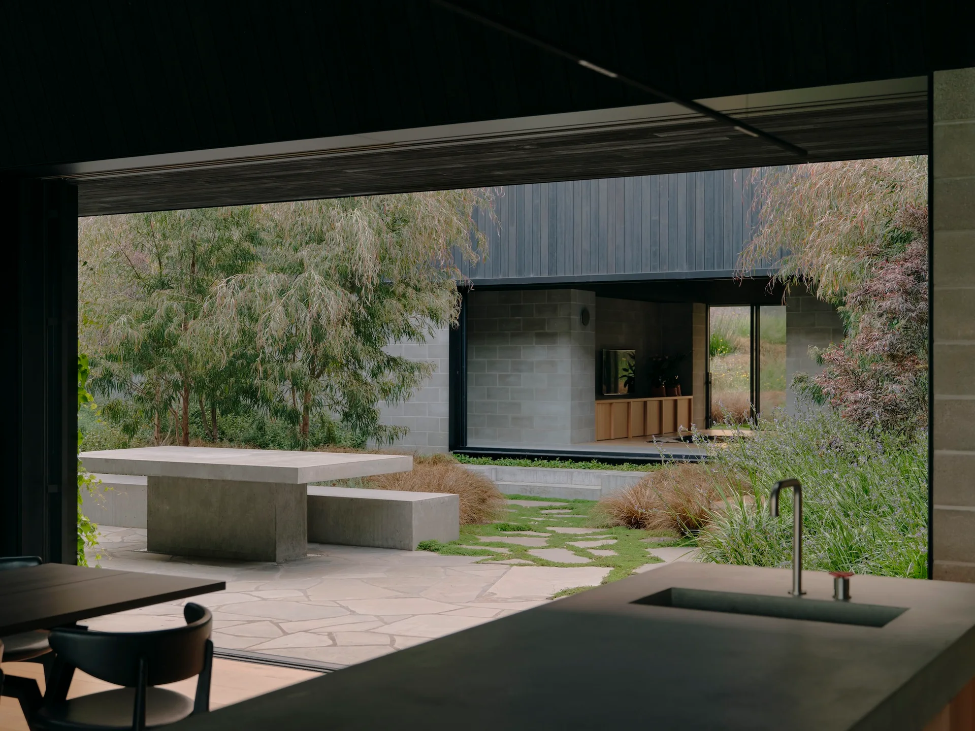 View from a dark kitchen counter looking out to a modern flagstone patio featuring a concrete dining table and bench surrounded by green landscaping.